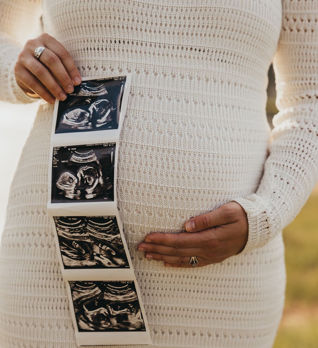 Pregnant woman in cream knit dress holding ultrasound photos against her baby bump.
