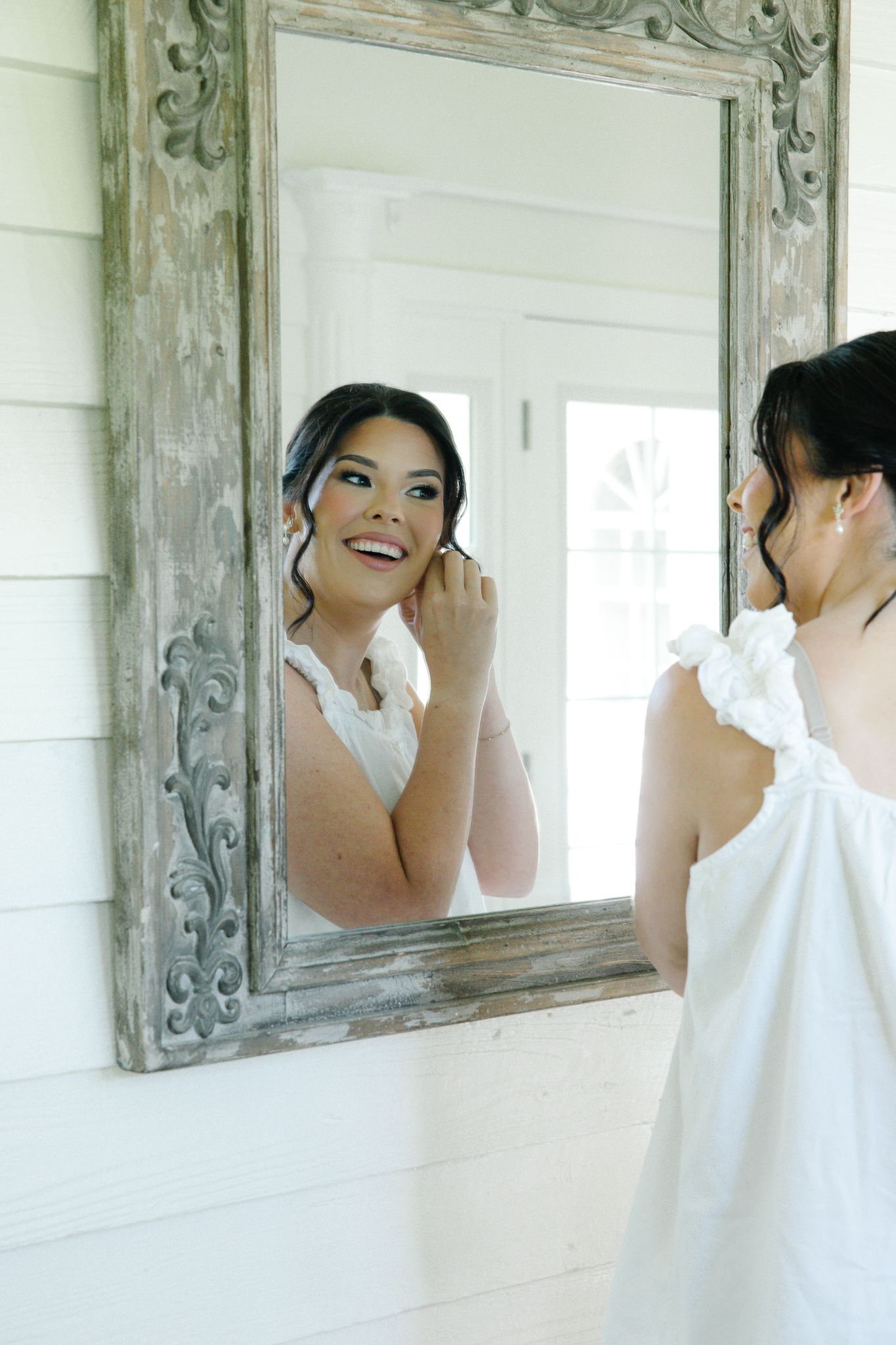 Smiling bride in white dress puts on earring while looking at her reflection in an ornate mirror.