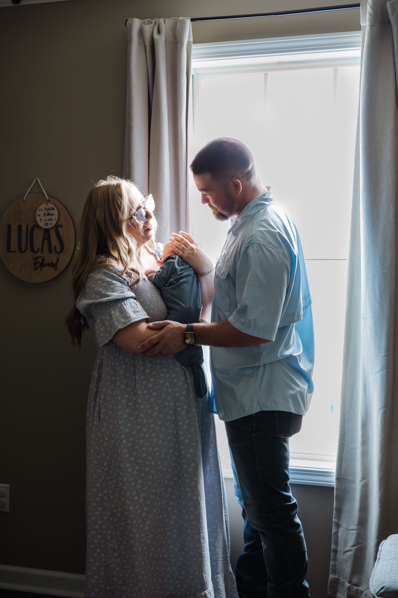 Parents stand by a bright window in a nursery, lovingly gazing at their newborn baby boy Lucas.