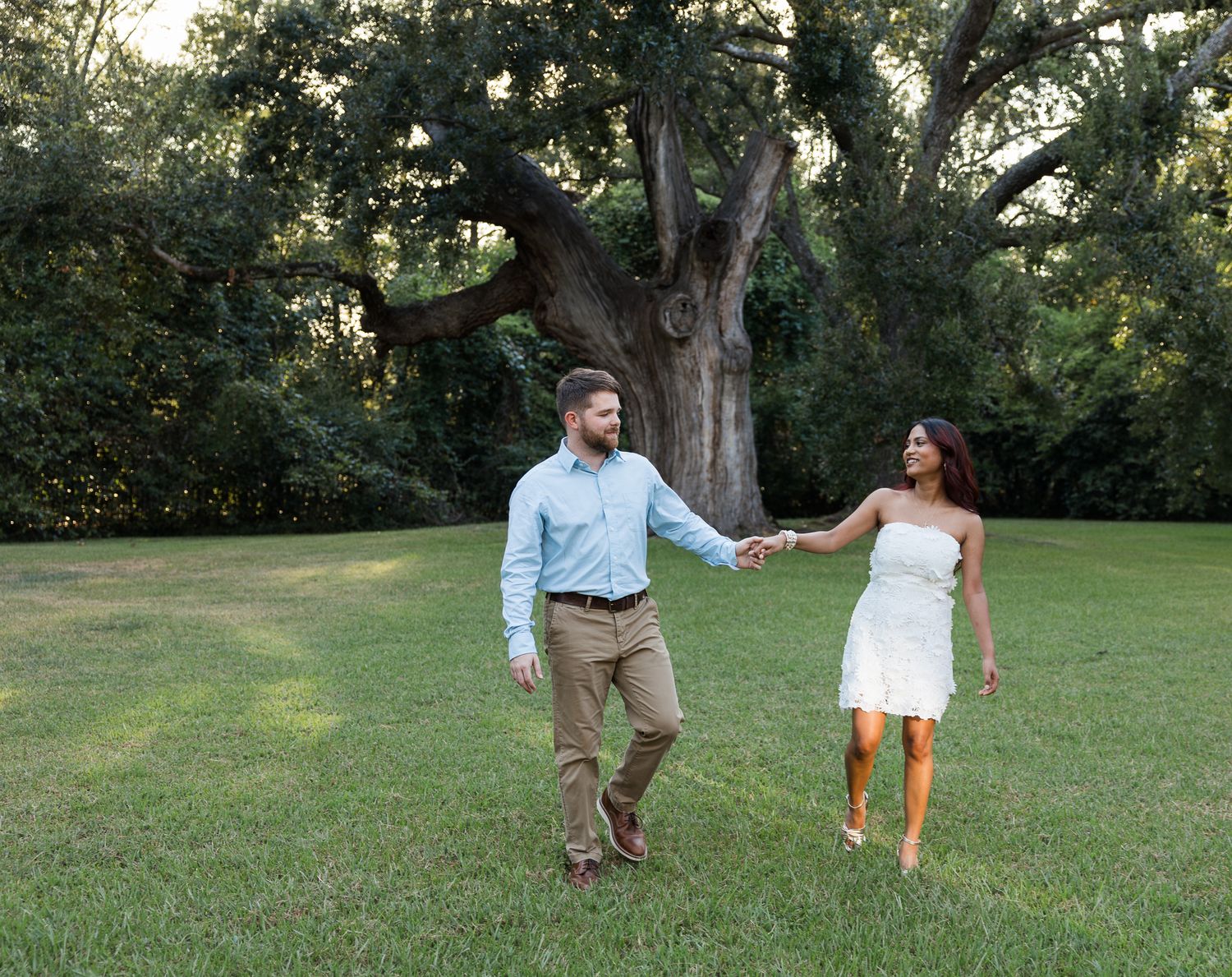 Couple holding hands walking on green grass with large oak tree backdrop in outdoor engagement photo.