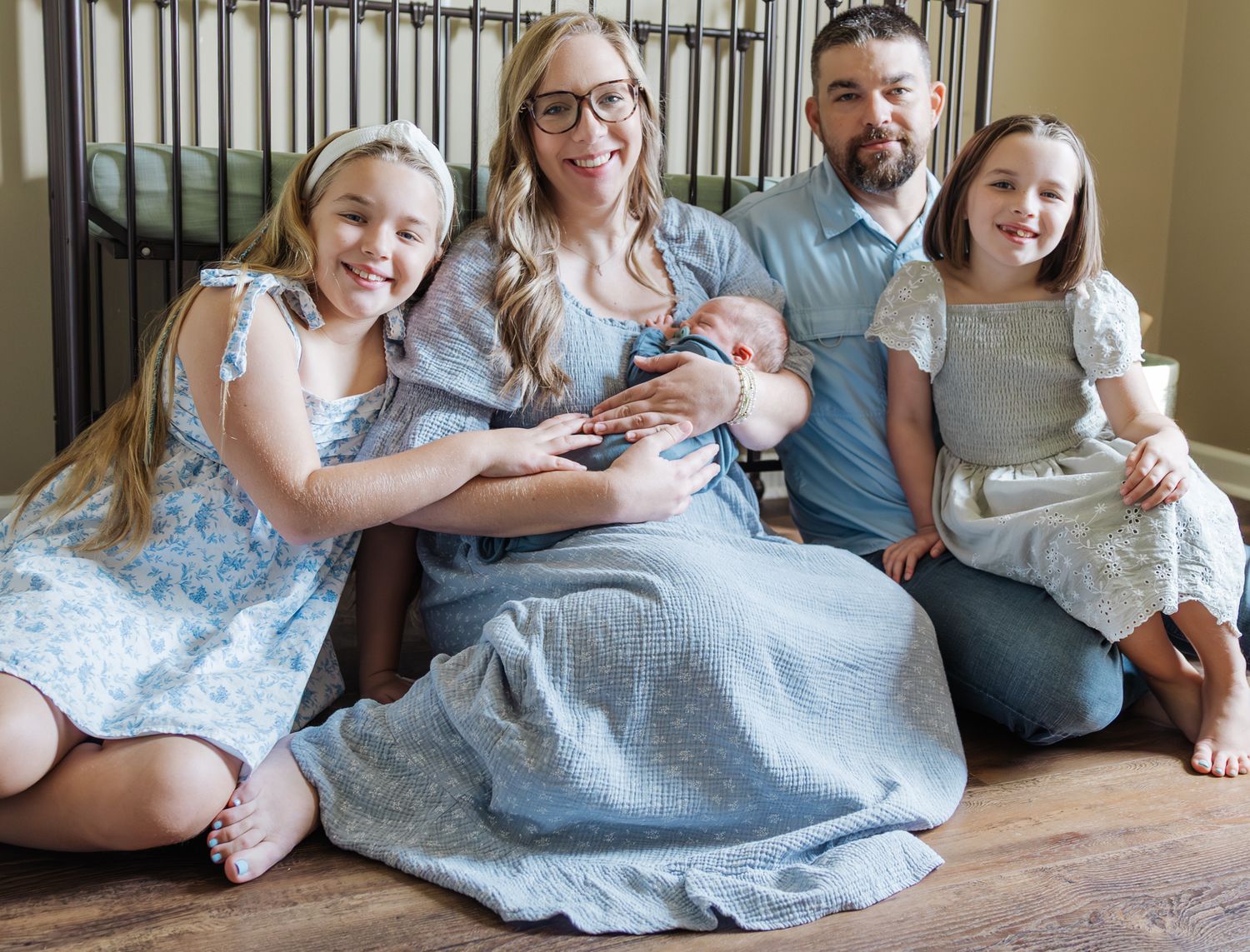 Happy family of five poses together with newborn baby boy sitting on nursery floor.
