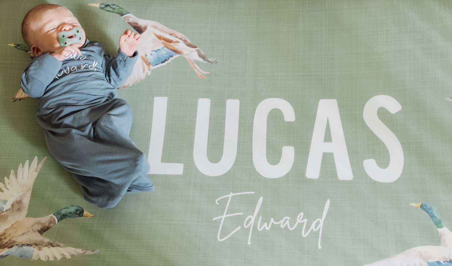 Newborn baby Lucas Edward lying on a green personalized name blanket in a blue onesie.