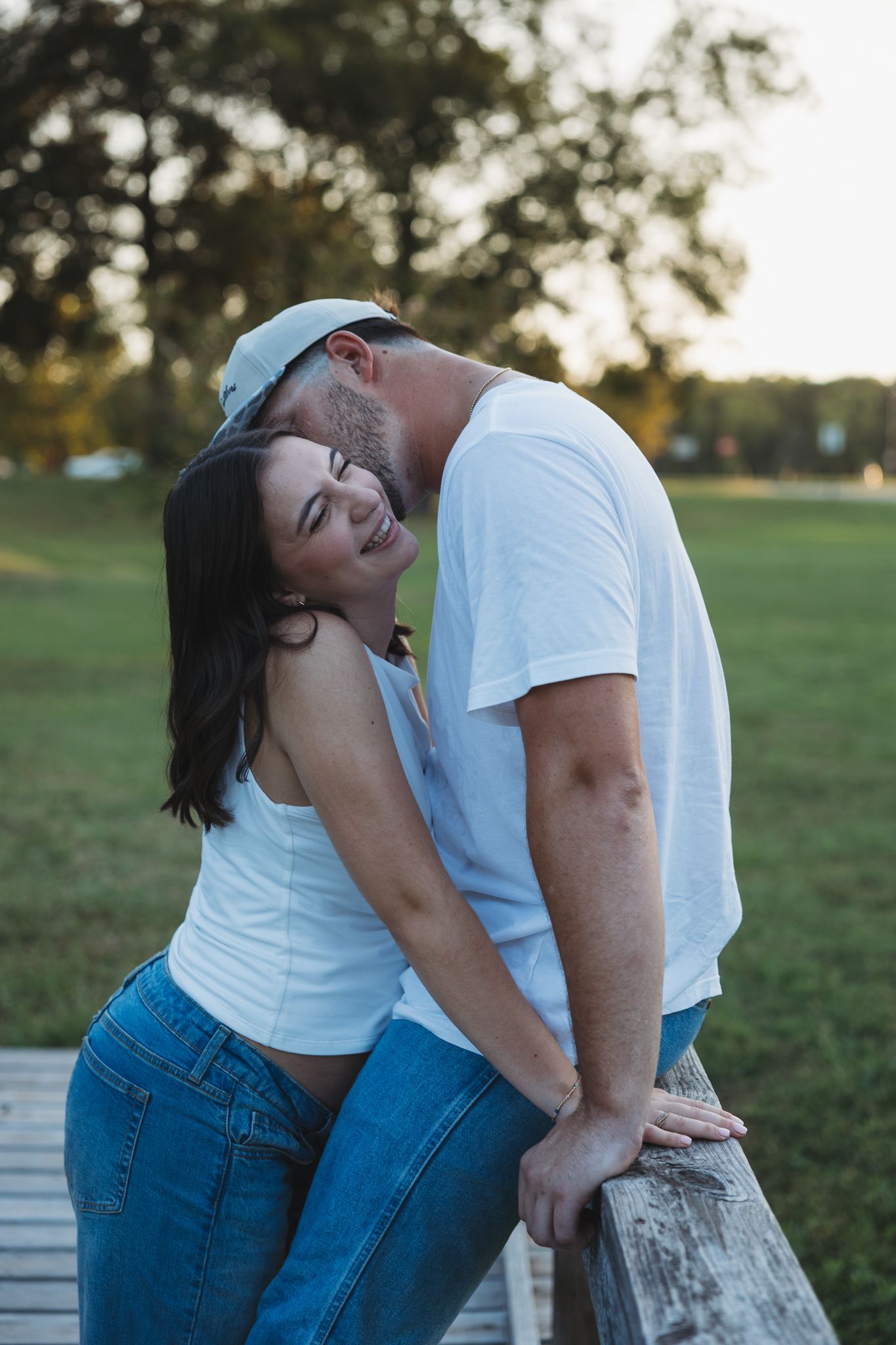 Happy couple laughing together leaning on wooden fence in park at golden hour, casual white and denim outfits.