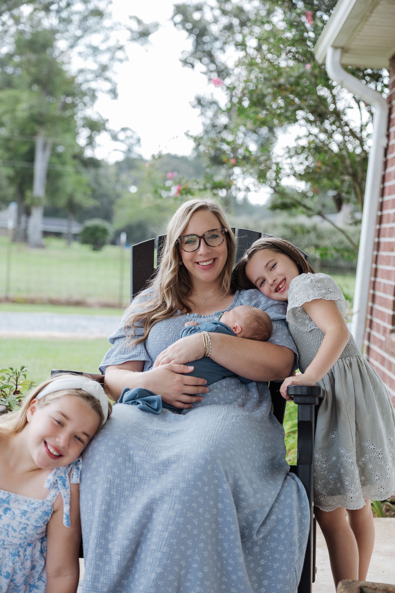 A mother holds her newborn while her two daughters lean against her in a rocking chair on a brick porch.