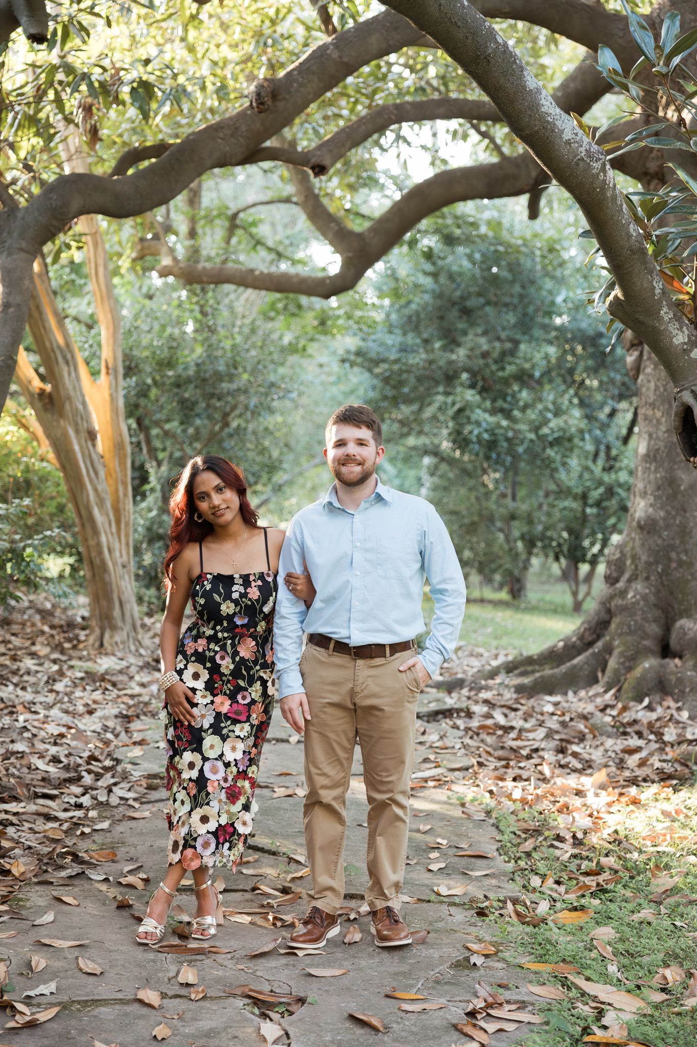 Couple posing together on a leaf-covered path surrounded by large trees in a lush green park setting.