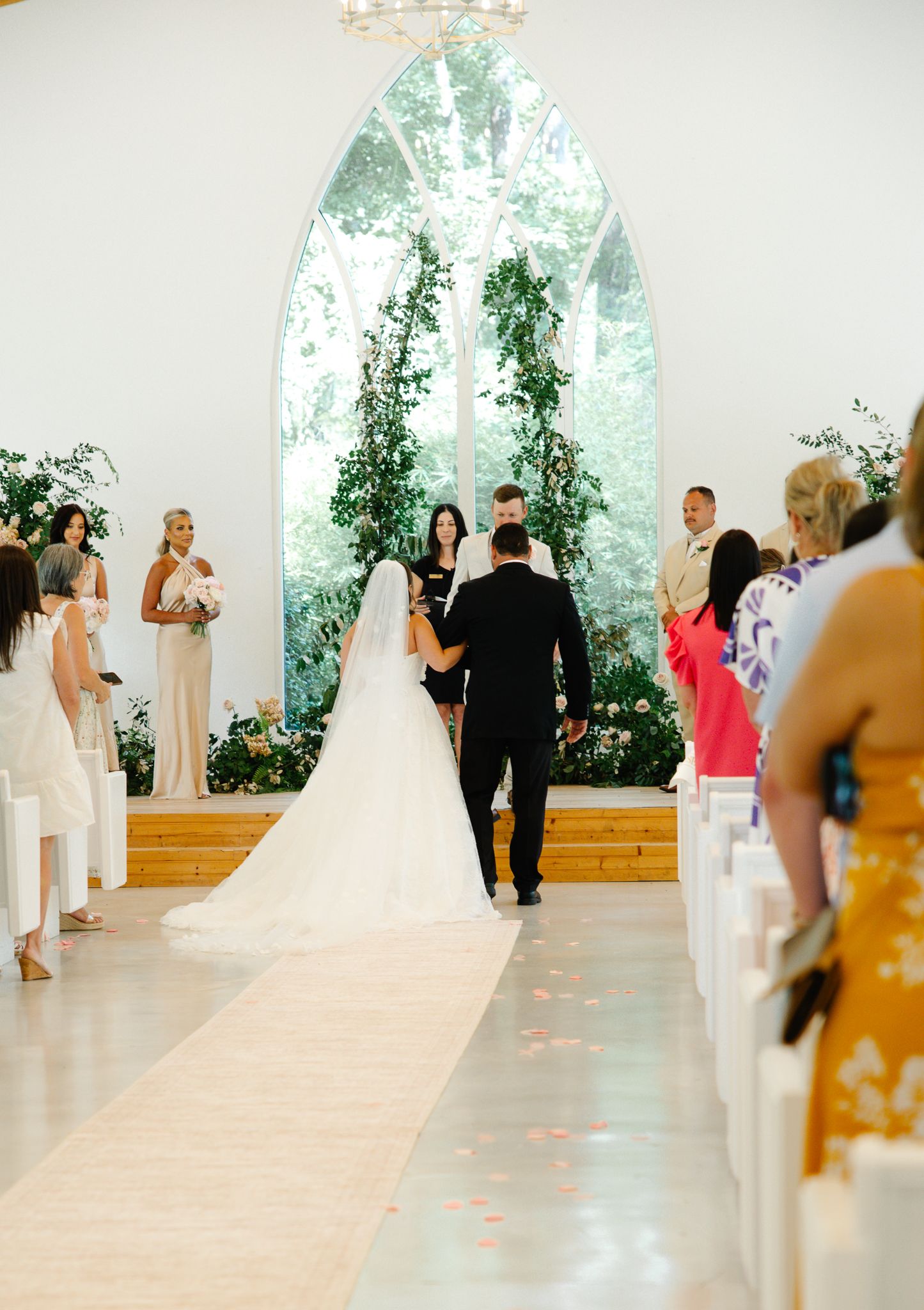 Bride in long veil walking down white chapel aisle toward groom during elegant wedding ceremony.