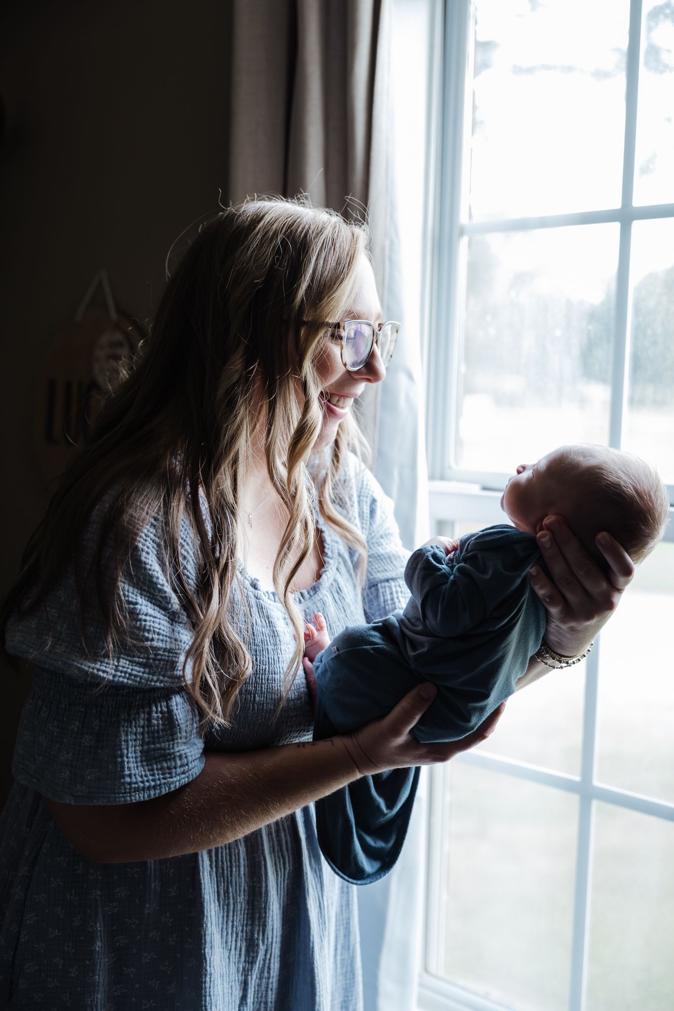 Smiling mother with glasses holding newborn baby near bright window in soft natural light.