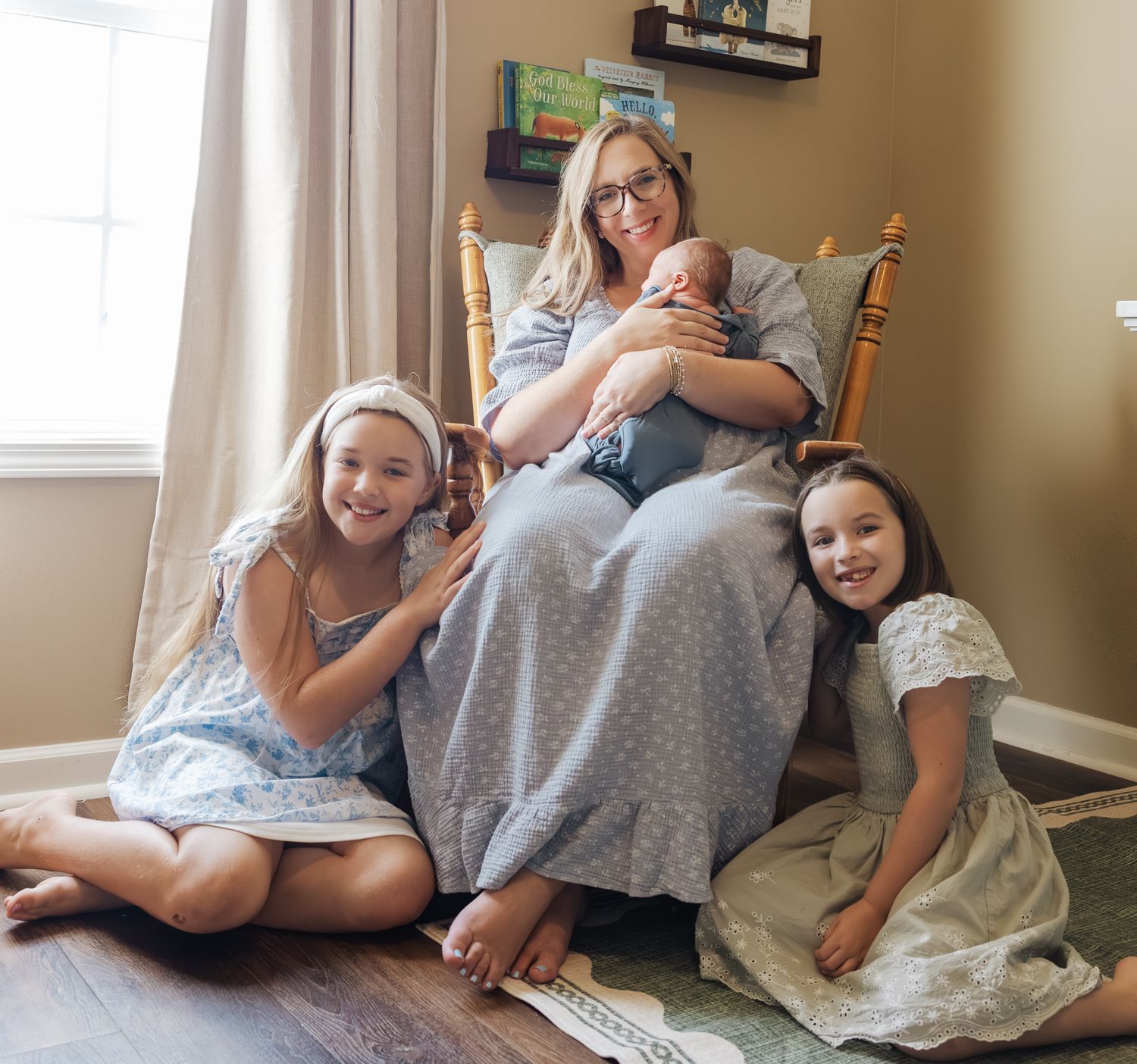 A mother sits in a rocking chair holding her newborn while her two young daughters smile beside her.