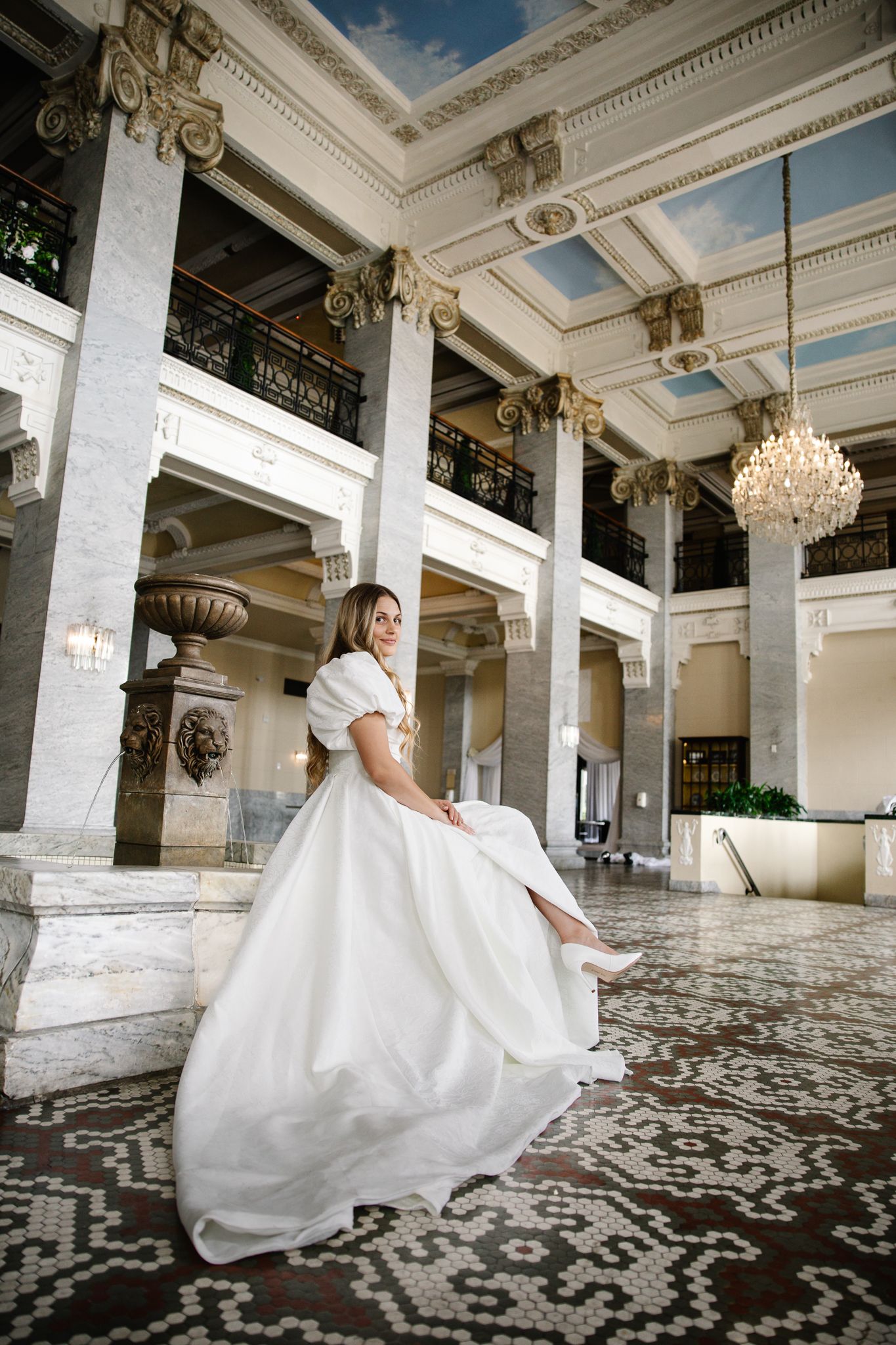 Elegant bride in a white ball gown sits gracefully in a grand marble hall with ornate columns and chandeliers.