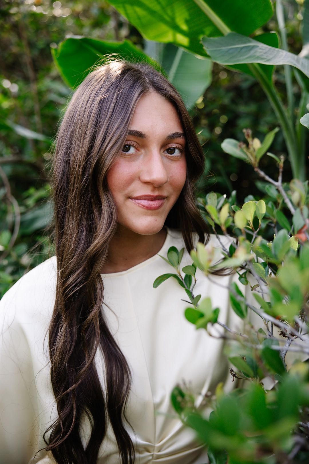 Young woman with long dark hair smiling softly among tropical green foliage and plants.