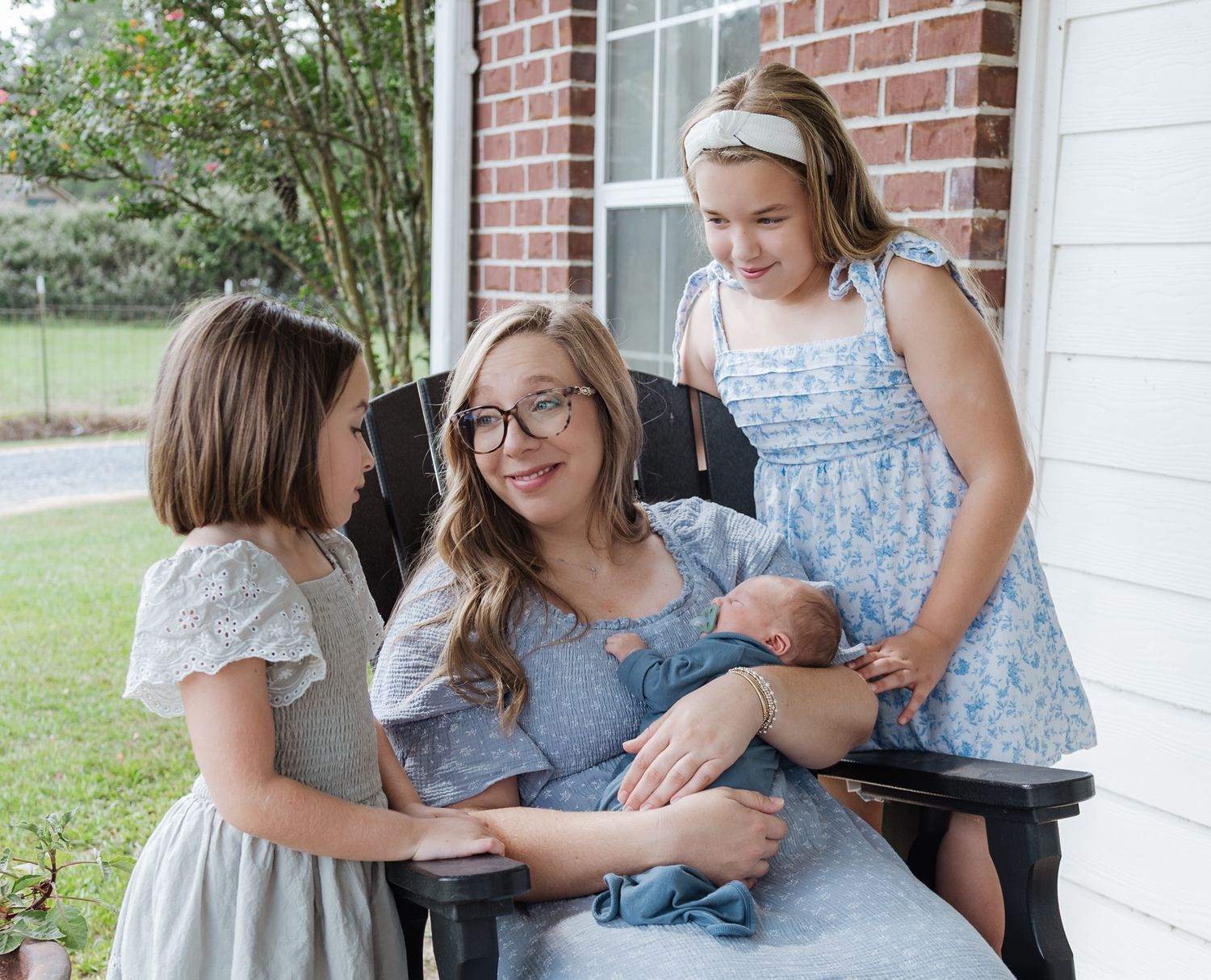 A smiling mother with glasses holds newborn baby Lucas on a porch surrounded by her two young daughters.