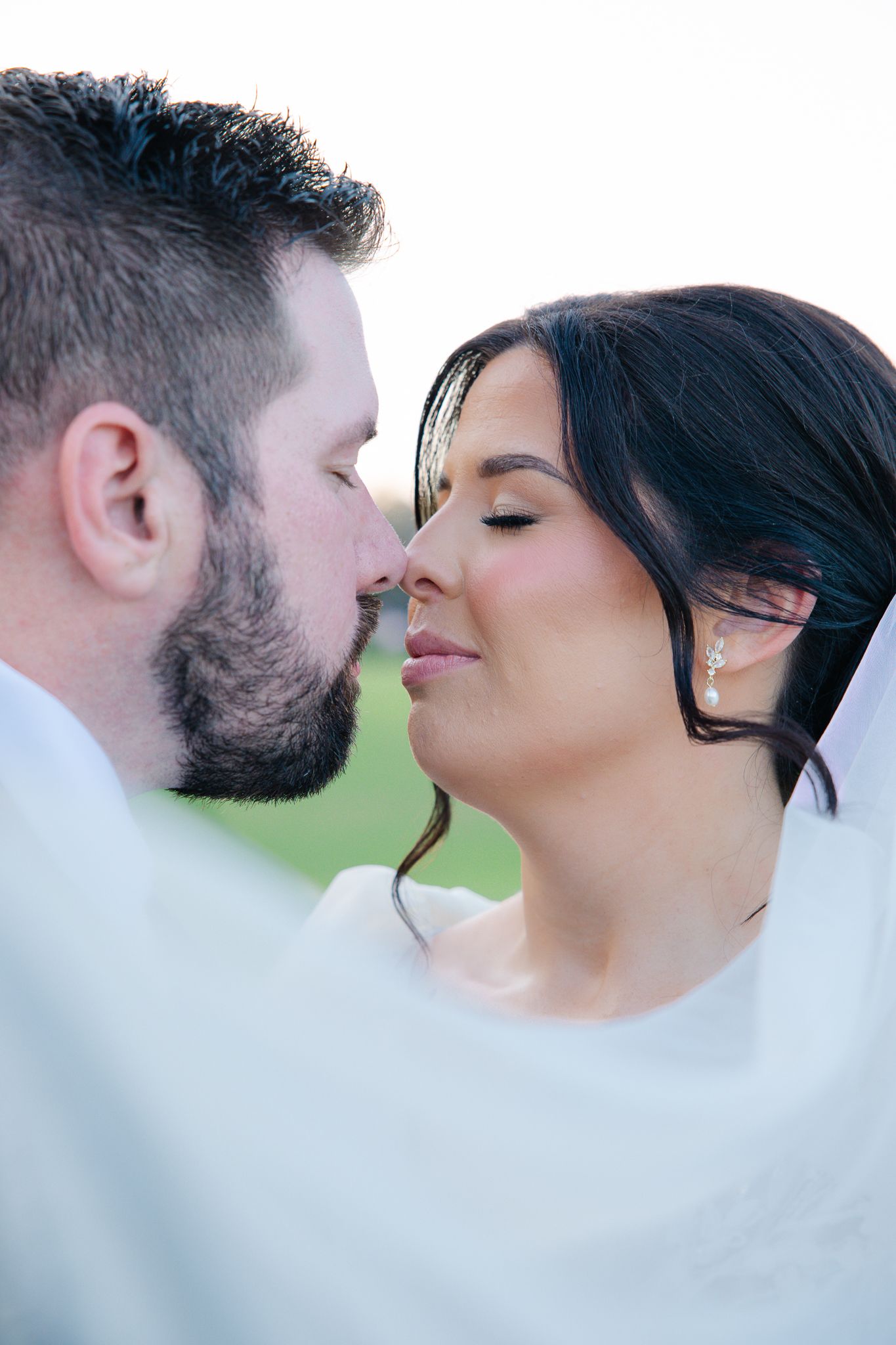 Bride and groom share a close romantic kiss outdoors wrapped in a flowing white veil.