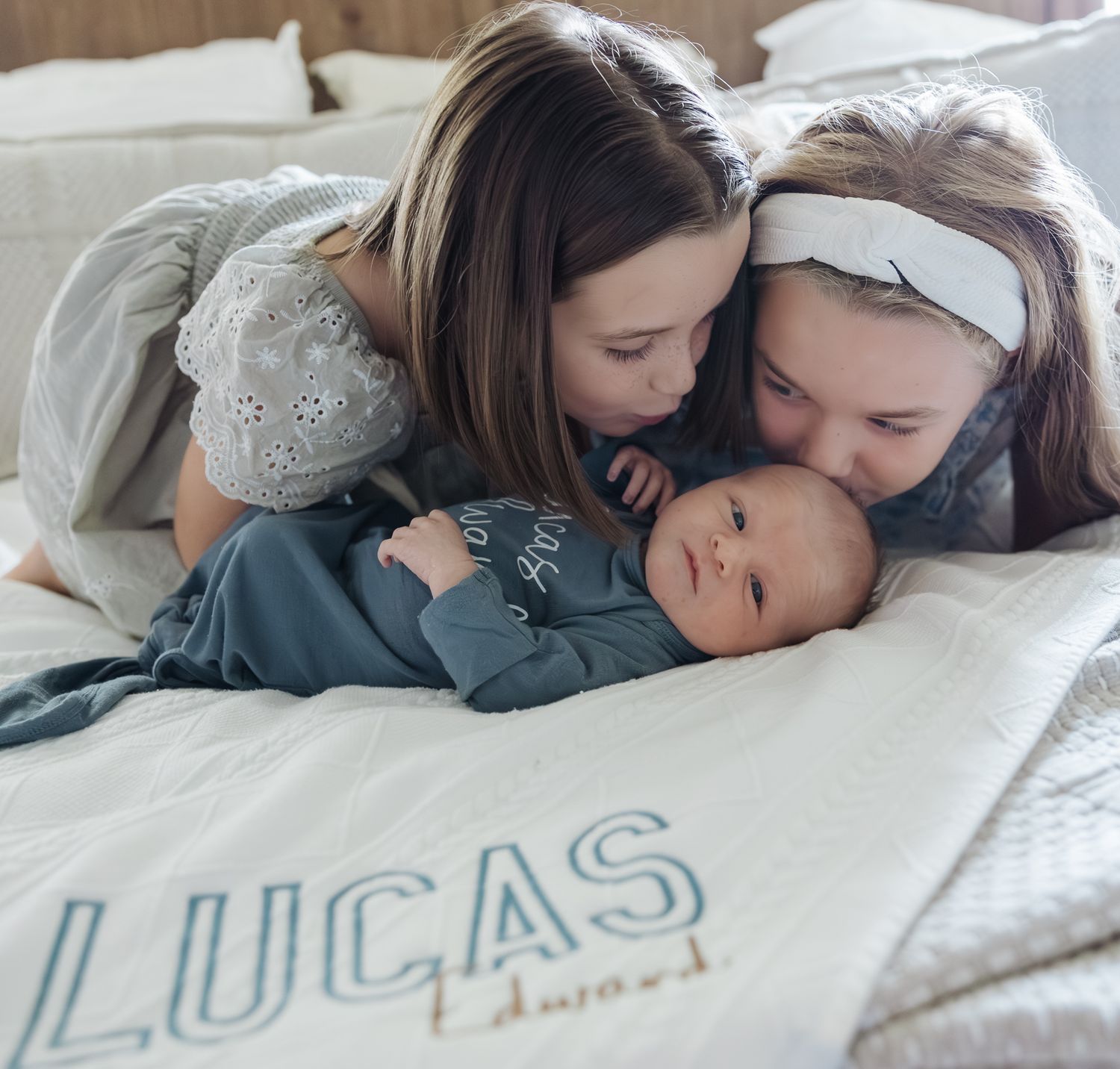 Two sisters kiss their newborn baby brother Lucas Edward on a white personalized blanket on a bed.