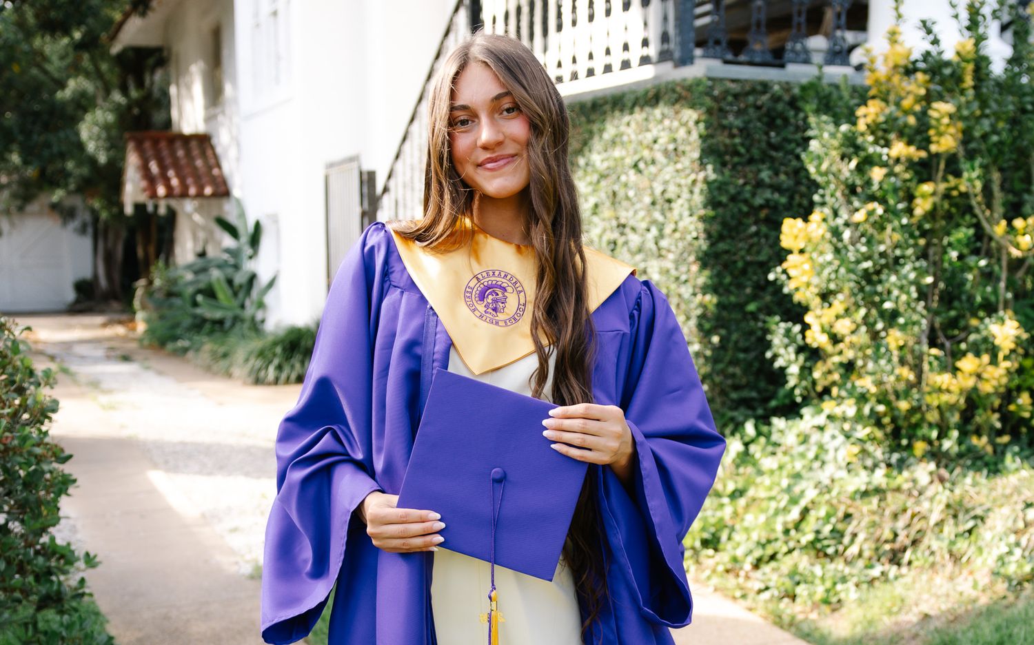Smiling graduate in purple cap and gown holding diploma outdoors in sunny garden setting.