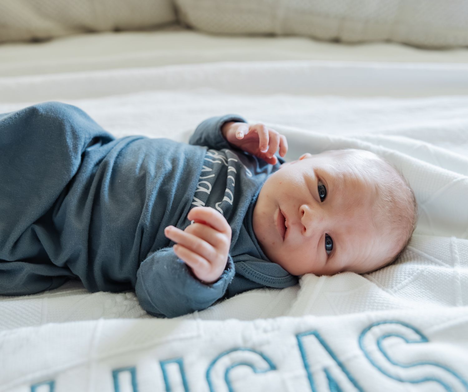 Alert newborn baby boy in blue outfit lying on white blanket with Lucas name embroidery.