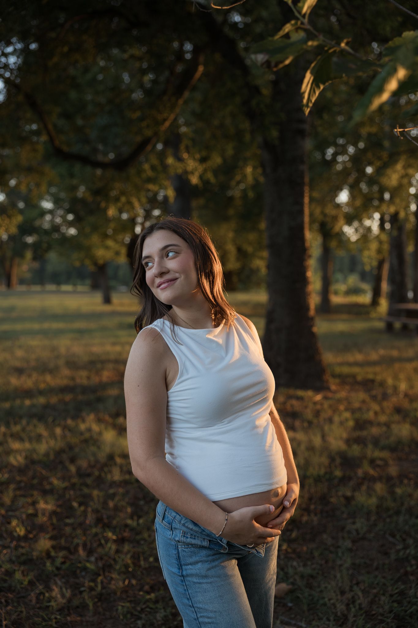 Pregnant woman in white tank top and jeans cradling her baby bump in a sunlit park during golden hour.