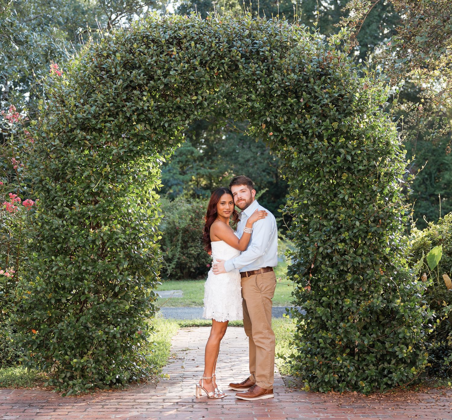 Couple embracing under a lush green garden arch, woman in white dress, man in casual attire, outdoor engagement photo.