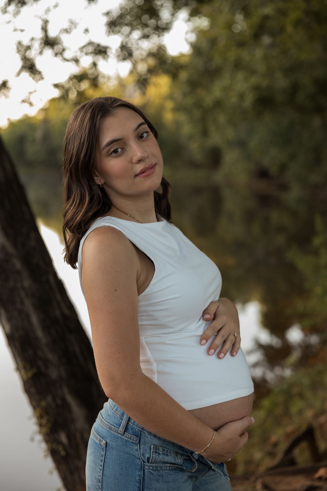 Pregnant woman in white sleeveless top and jeans cradling baby bump near tree by lake at sunset.