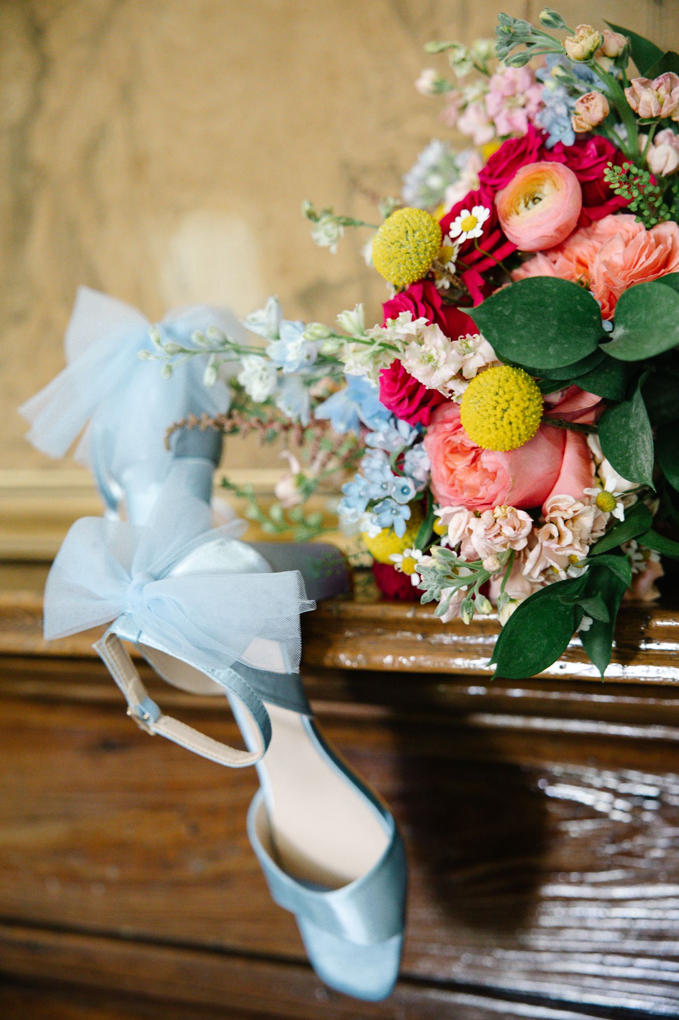 Light blue satin bridal heels with bow detail rest beside a colorful wedding bouquet on a wooden surface.