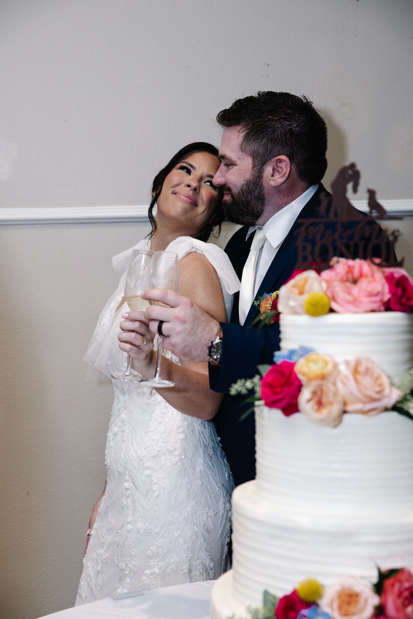 Bride and groom share a champagne toast beside a floral wedding cake at their reception.