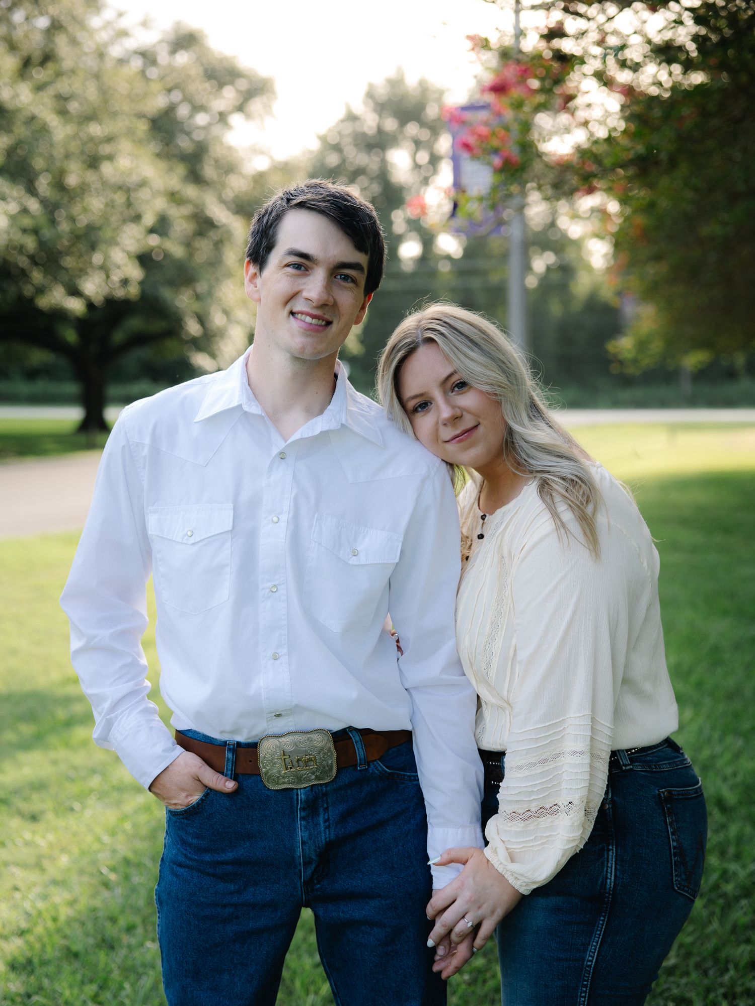 Smiling engaged couple posing together outdoors in park, man in white shirt and woman in cream top.