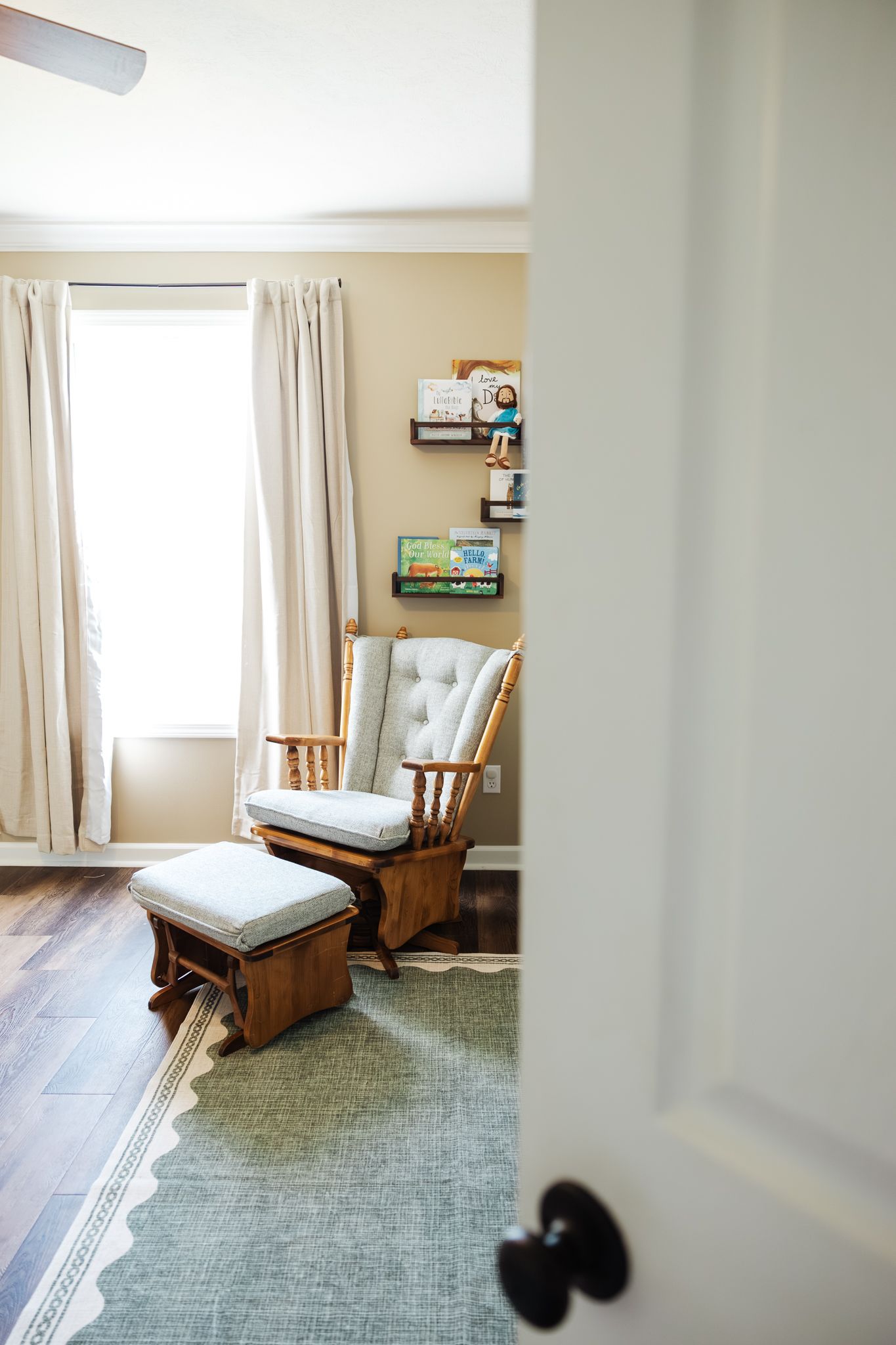 Bright nursery room with wooden glider rocking chair, ottoman, and book shelves on wall.