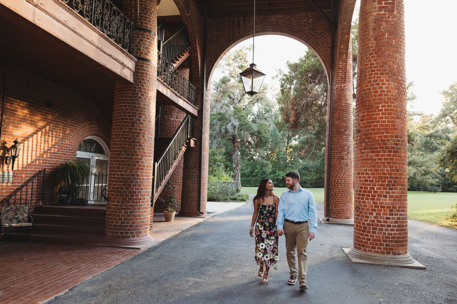 Couple walking hand-in-hand under brick arches of historic Southern mansion with grand staircase.