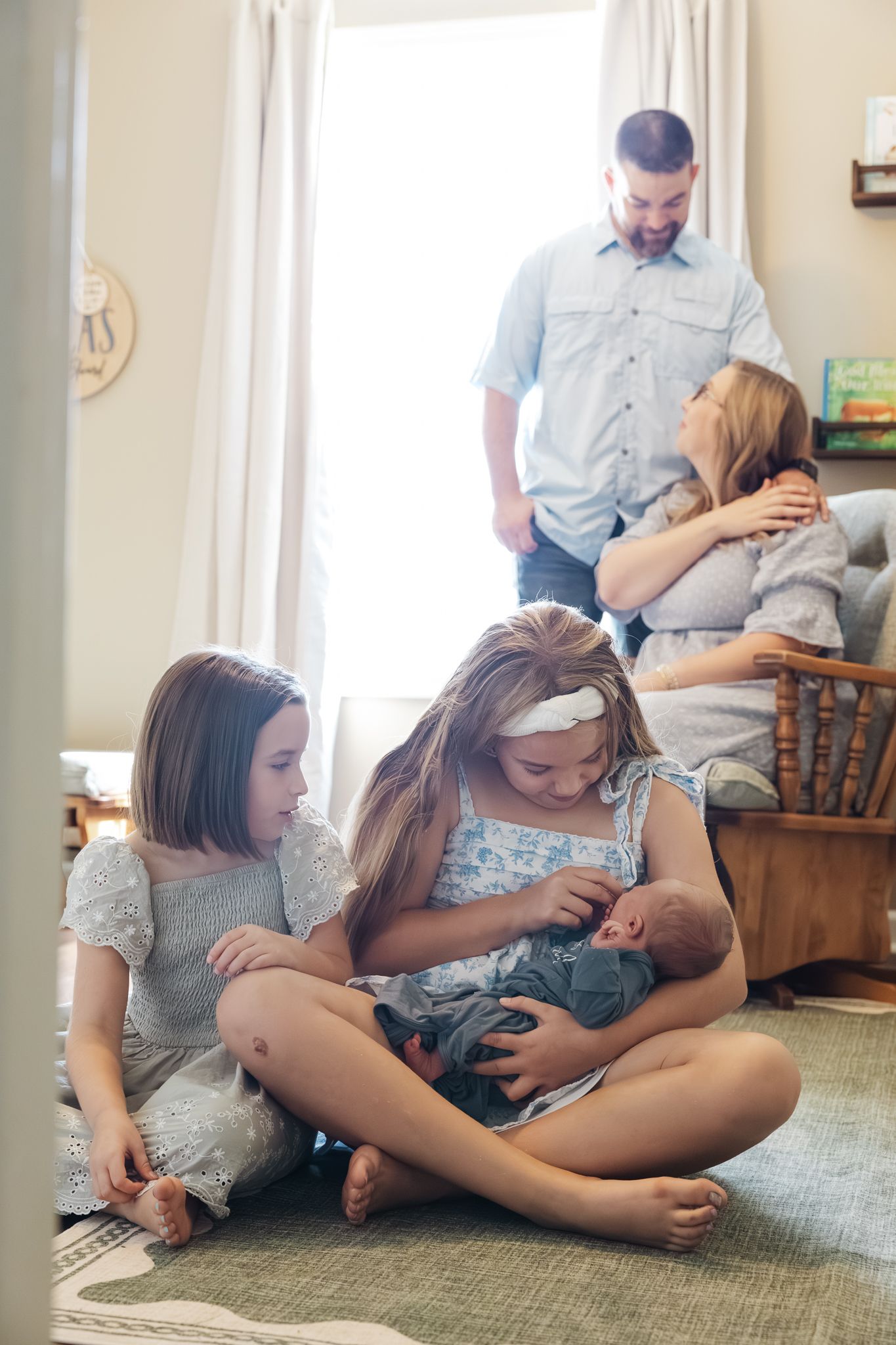 Two sisters sit on floor holding newborn while parents share tender moment behind them.