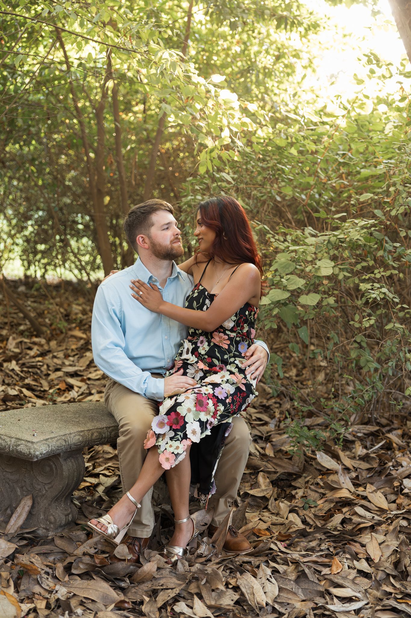 Couple sitting on stone bench surrounded by autumn leaves, woman in floral dress embracing man in blue shirt outdoors.