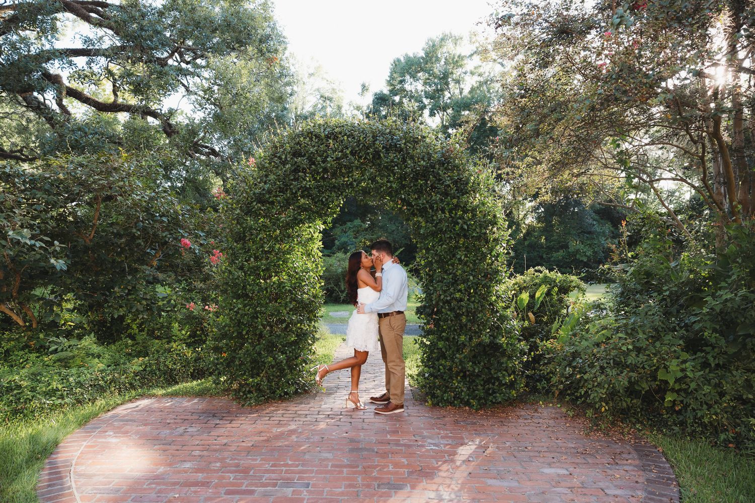 Romantic couple sharing a kiss under a lush green ivy archway on a brick pathway in a garden.