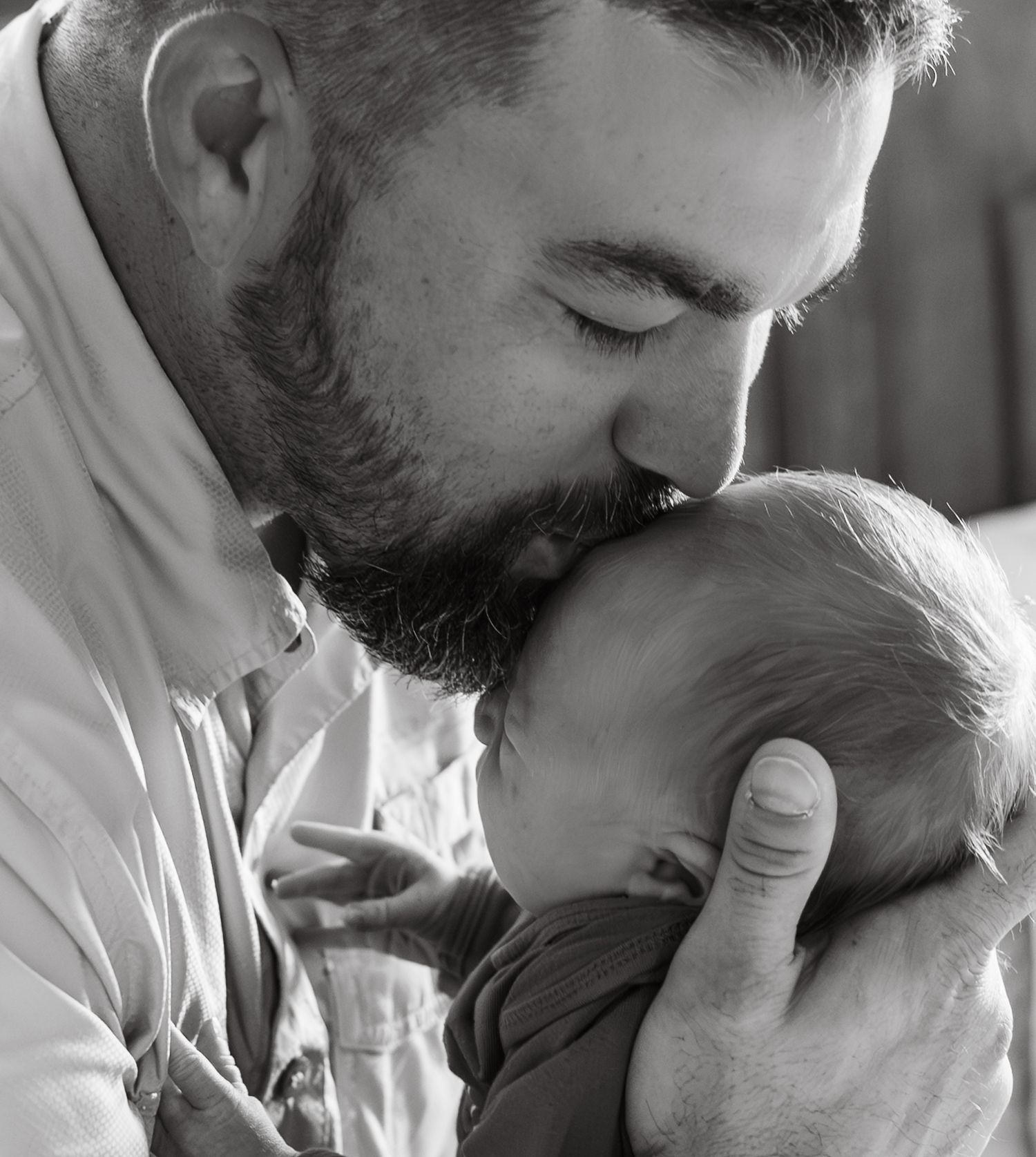 Bearded father tenderly kissing his newborn baby's head in a black and white portrait.