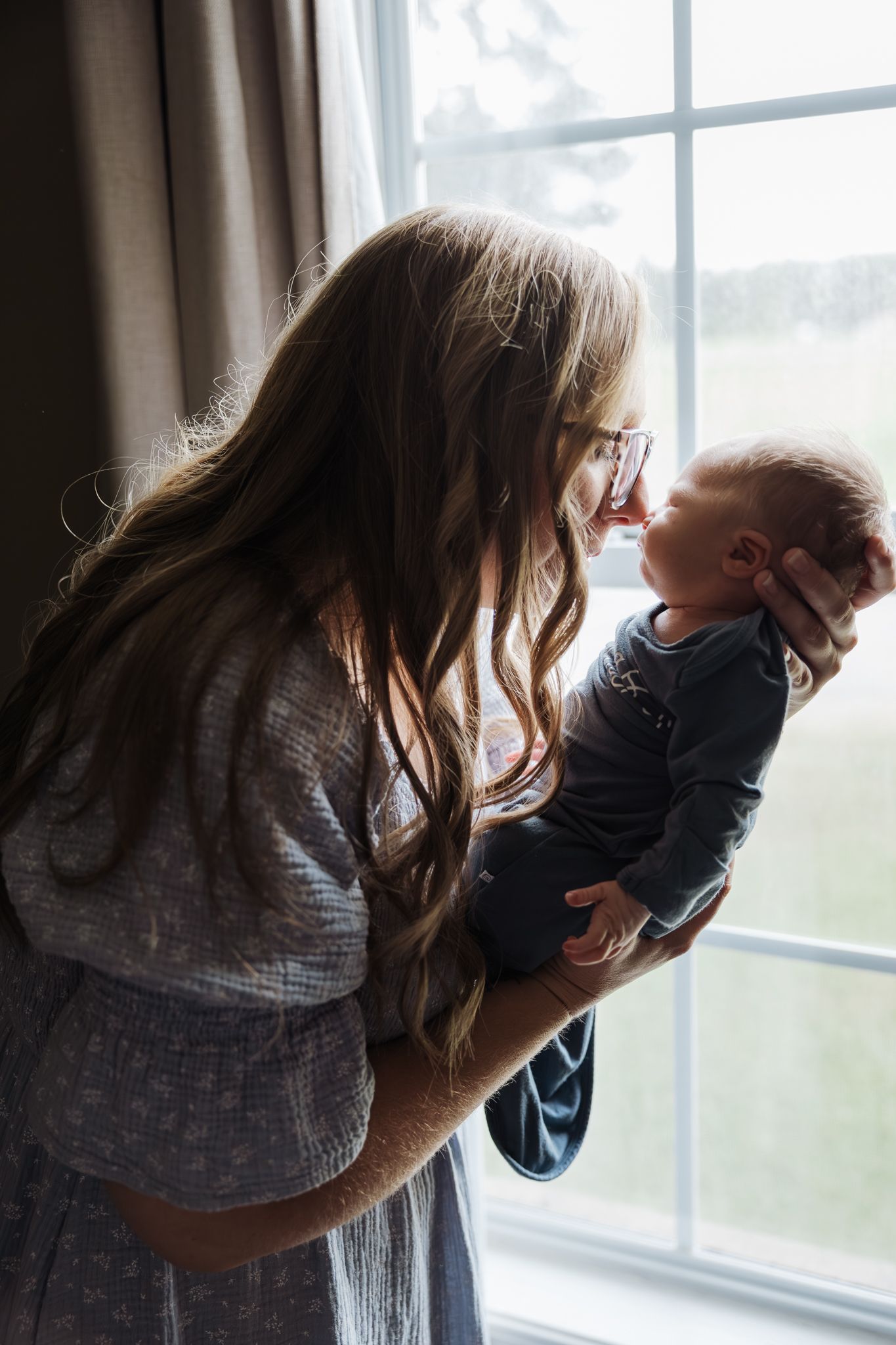 A long-haired mother with glasses holds her newborn baby up to her face near a large window.