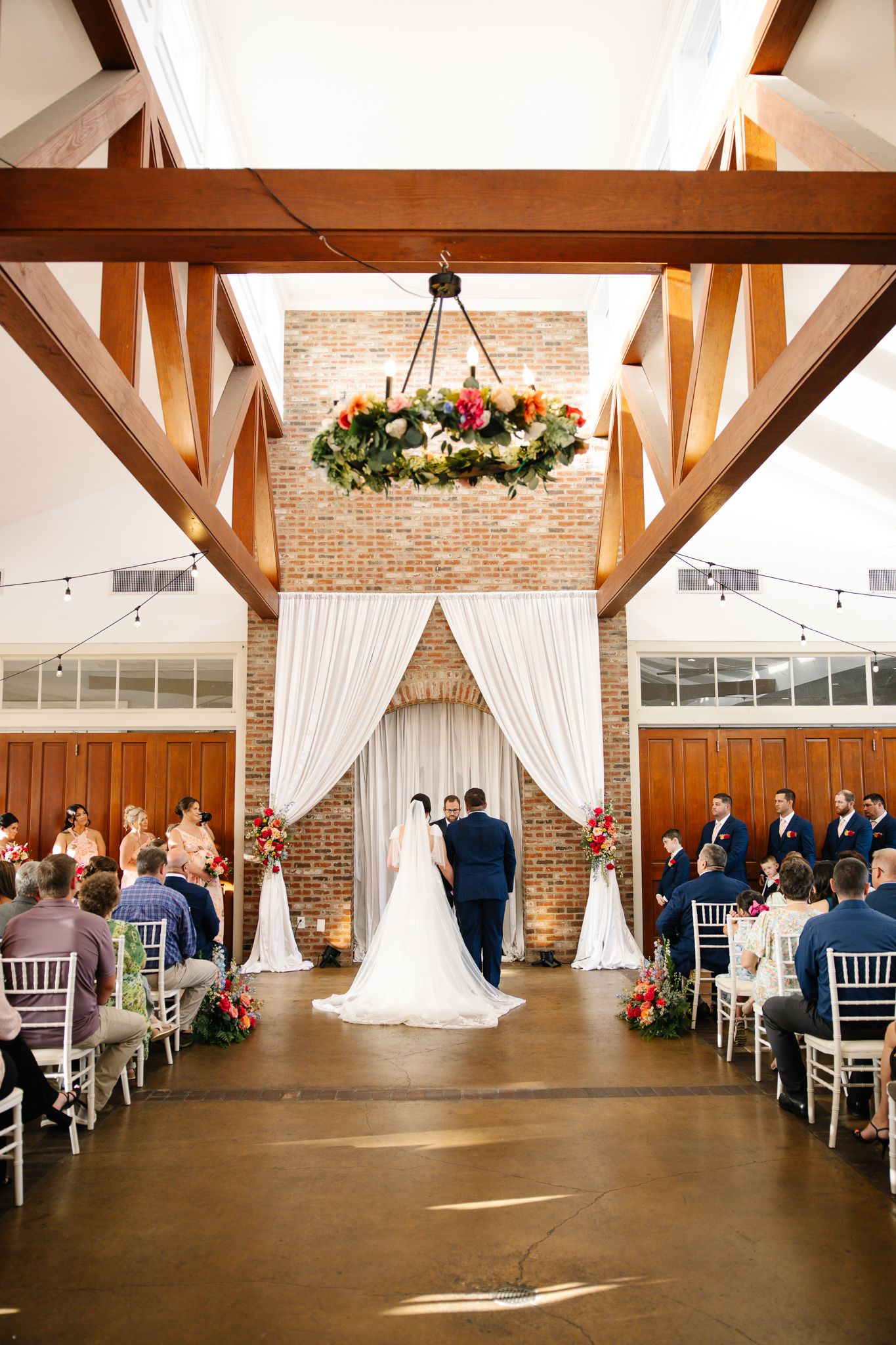 Wedding ceremony inside rustic venue with exposed brick, white drape arch, and floral chandelier overhead.