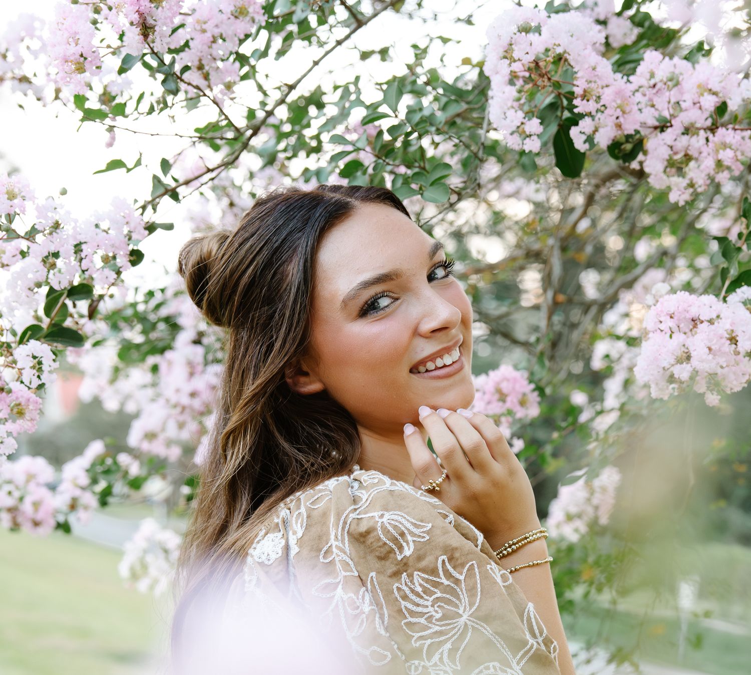 Young woman in floral lace dress smiling among blooming pink cherry blossom trees in a garden.