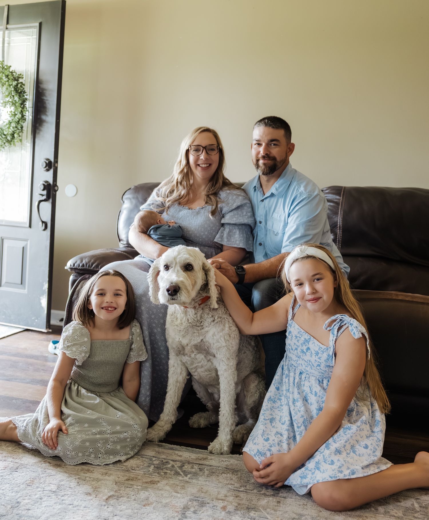 Family portrait with parents, two daughters, newborn baby, and white goldendoodle in cozy home.