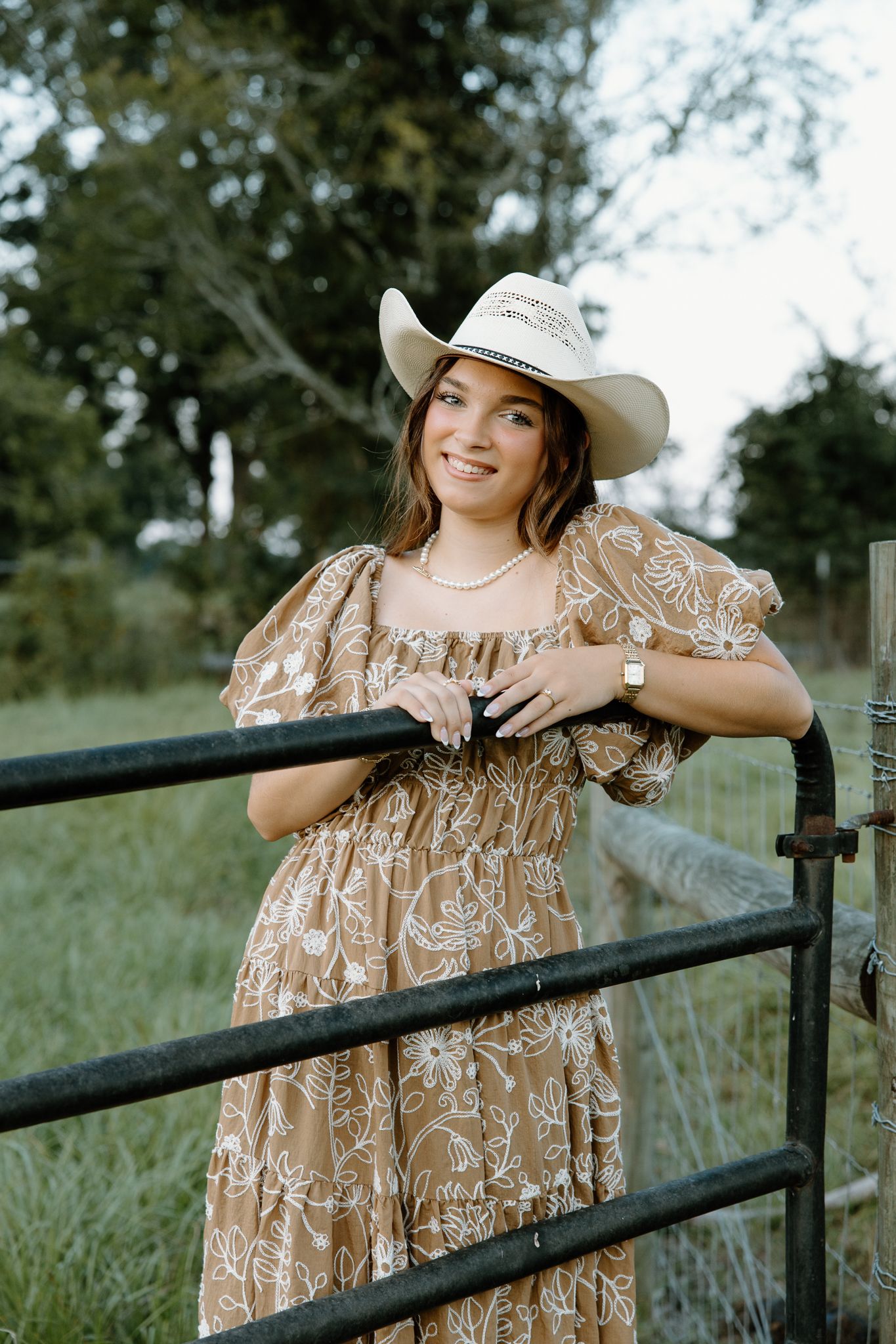Young woman in floral dress and white cowboy hat leaning on a farm gate, smiling outdoors.