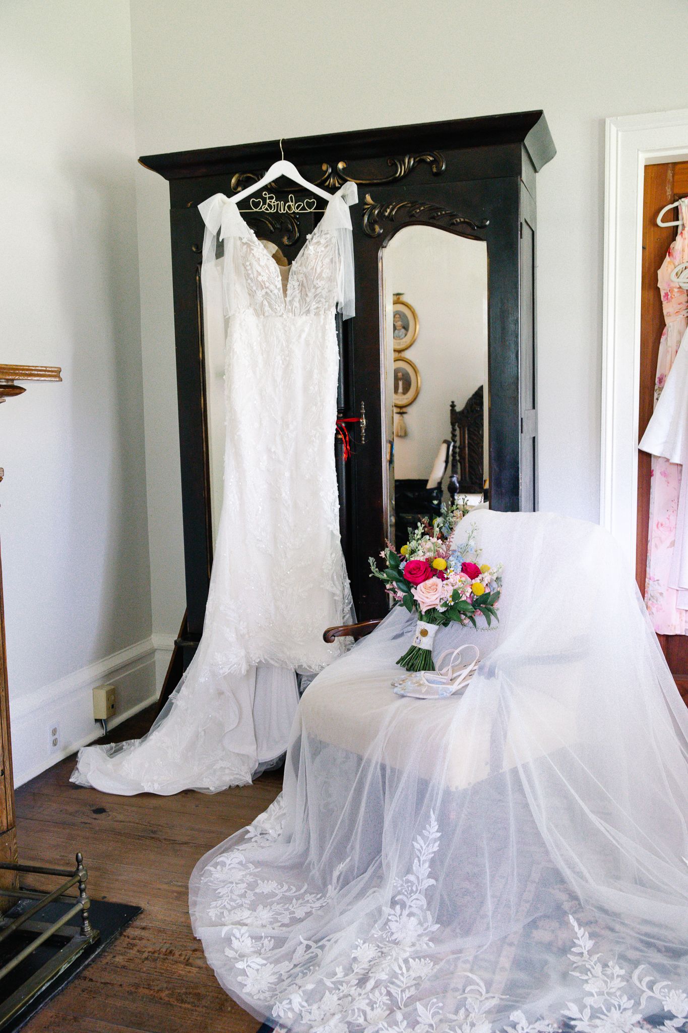 Elegant lace wedding dress hanging on an armoire with bridal bouquet and veil displayed nearby.