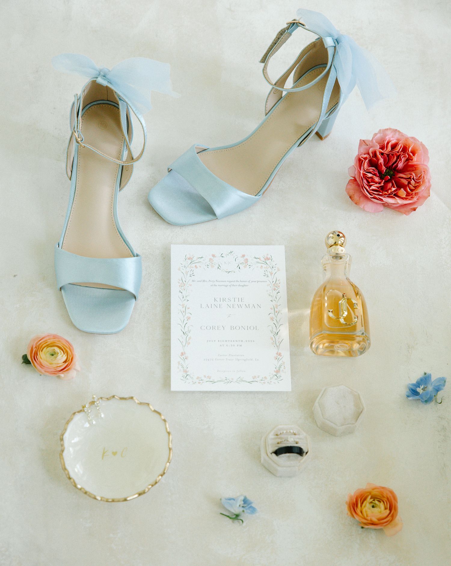 Flat lay of light blue bridal heels, wedding invitation, perfume, rings, and colorful ranunculus flowers.