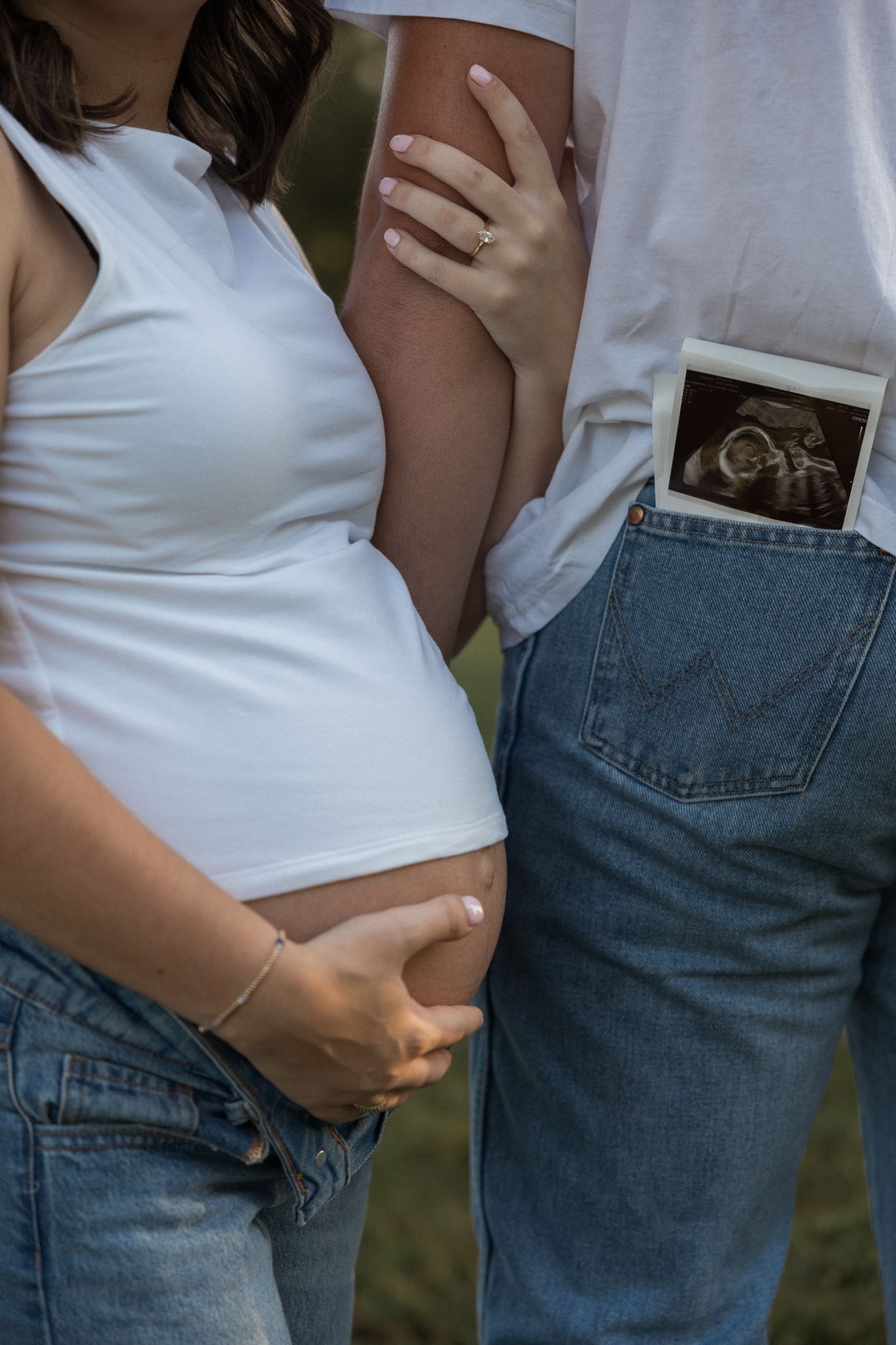 Pregnant woman in white tank top and jeans with partner holding ultrasound photo in back pocket outdoors.