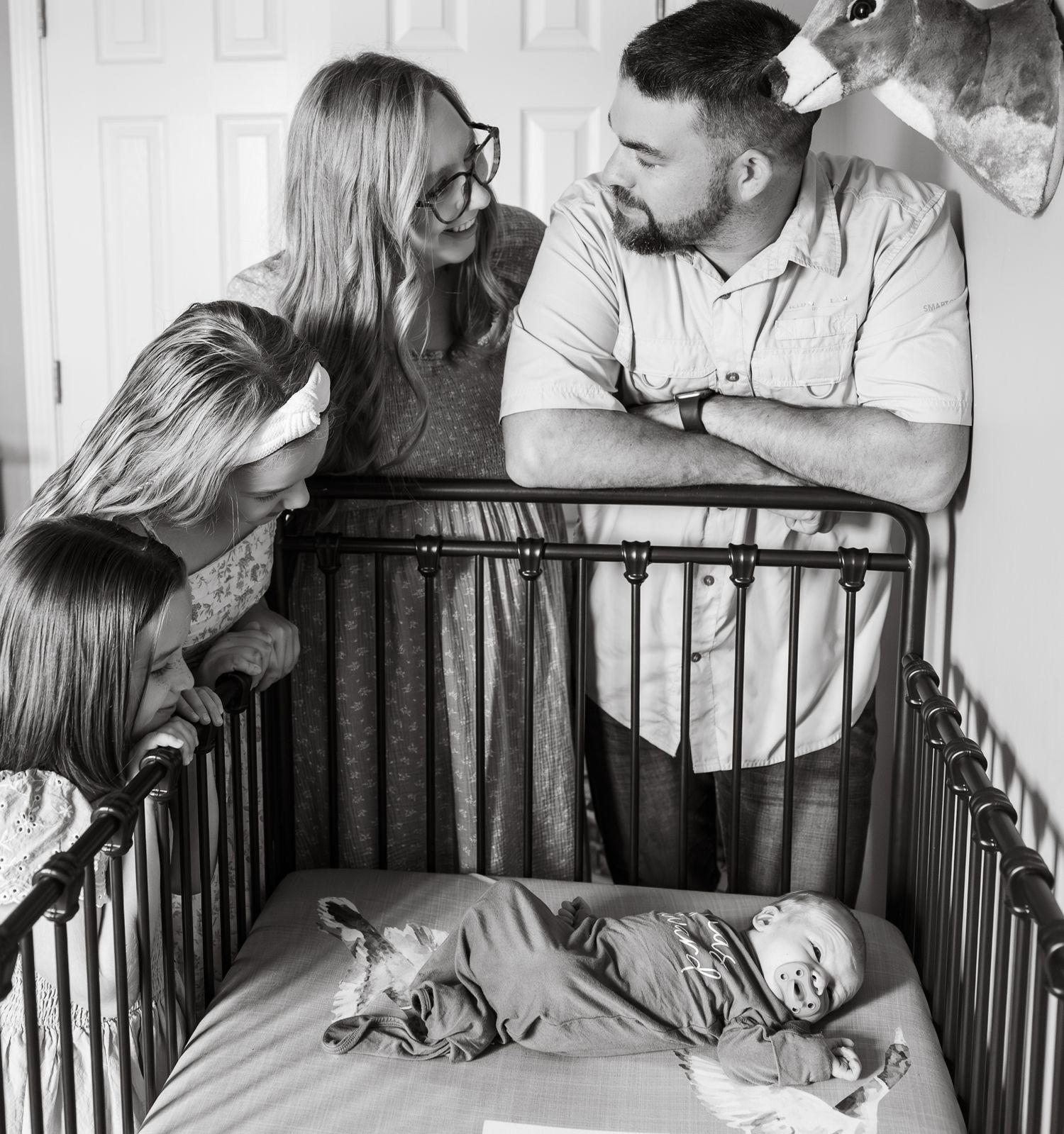 Parents and two children lovingly look down at a newborn baby lying in a crib, black and white photo.