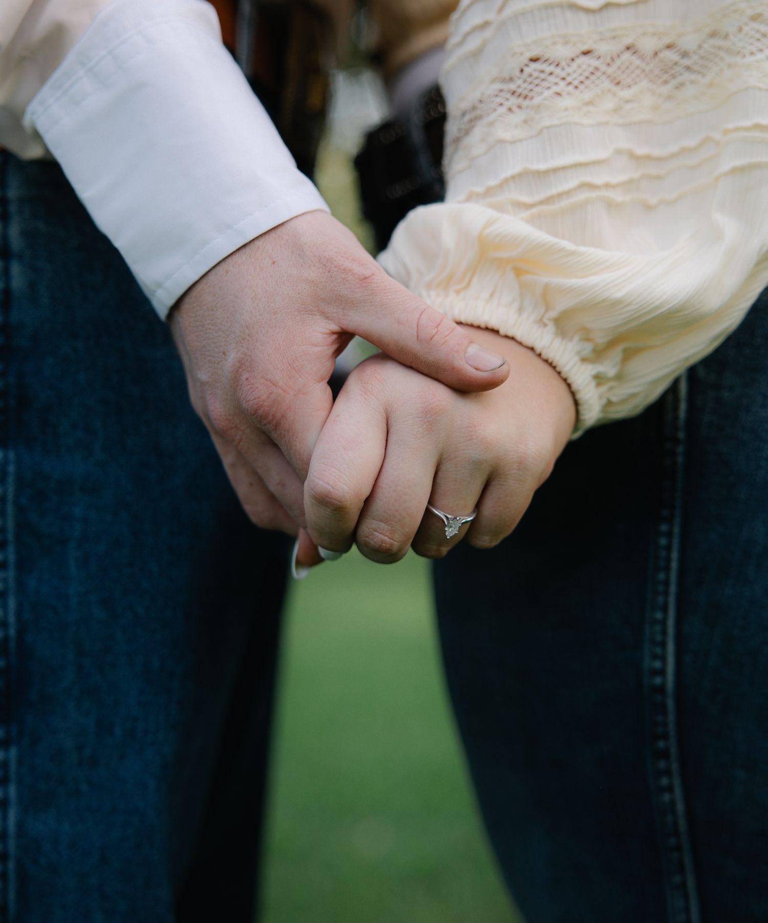 Close-up of couple holding hands with engagement ring visible, wearing jeans and cream top.