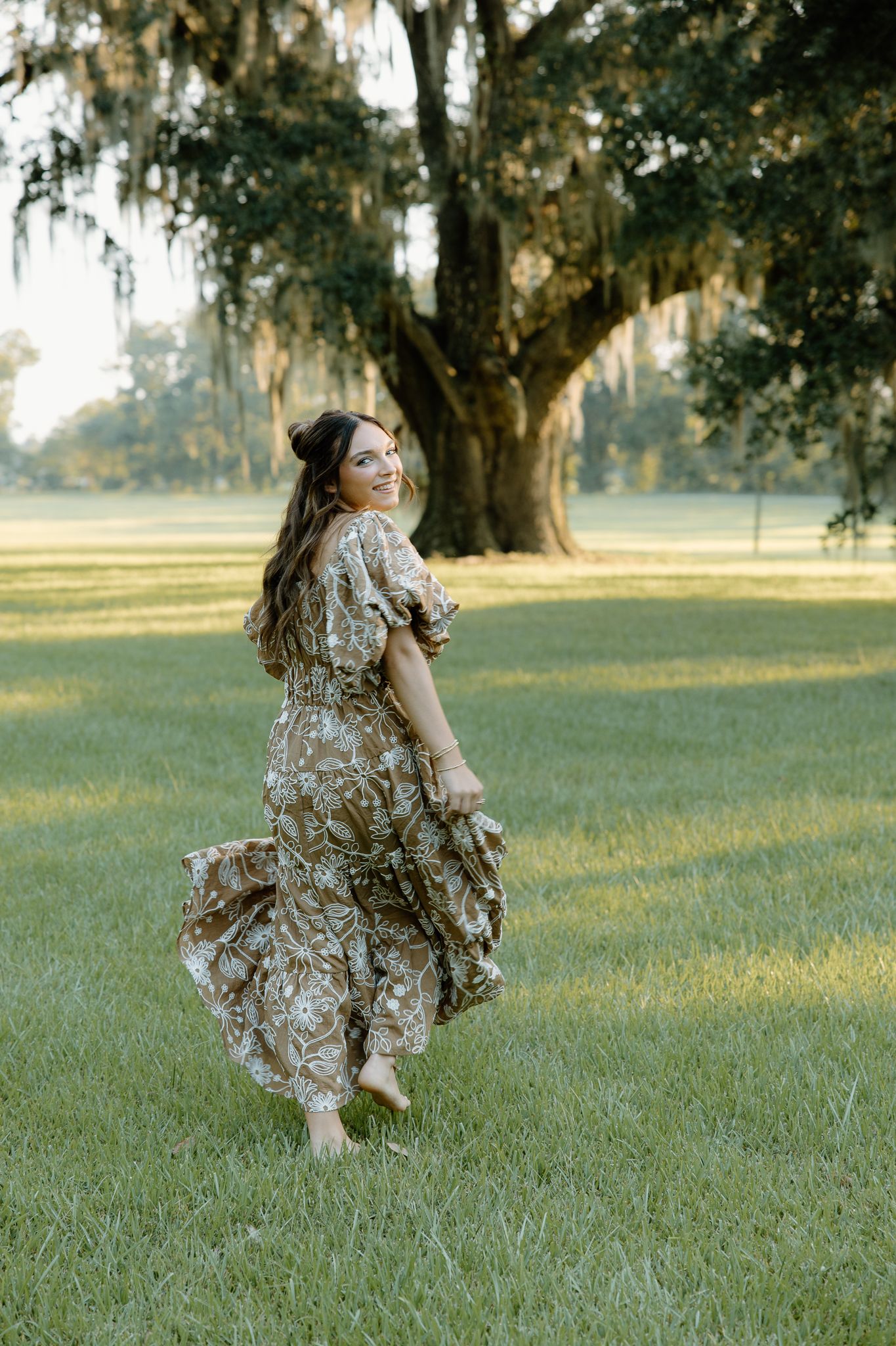 Woman in floral maxi dress twirling barefoot on green grass near a large moss-draped oak tree outdoors.