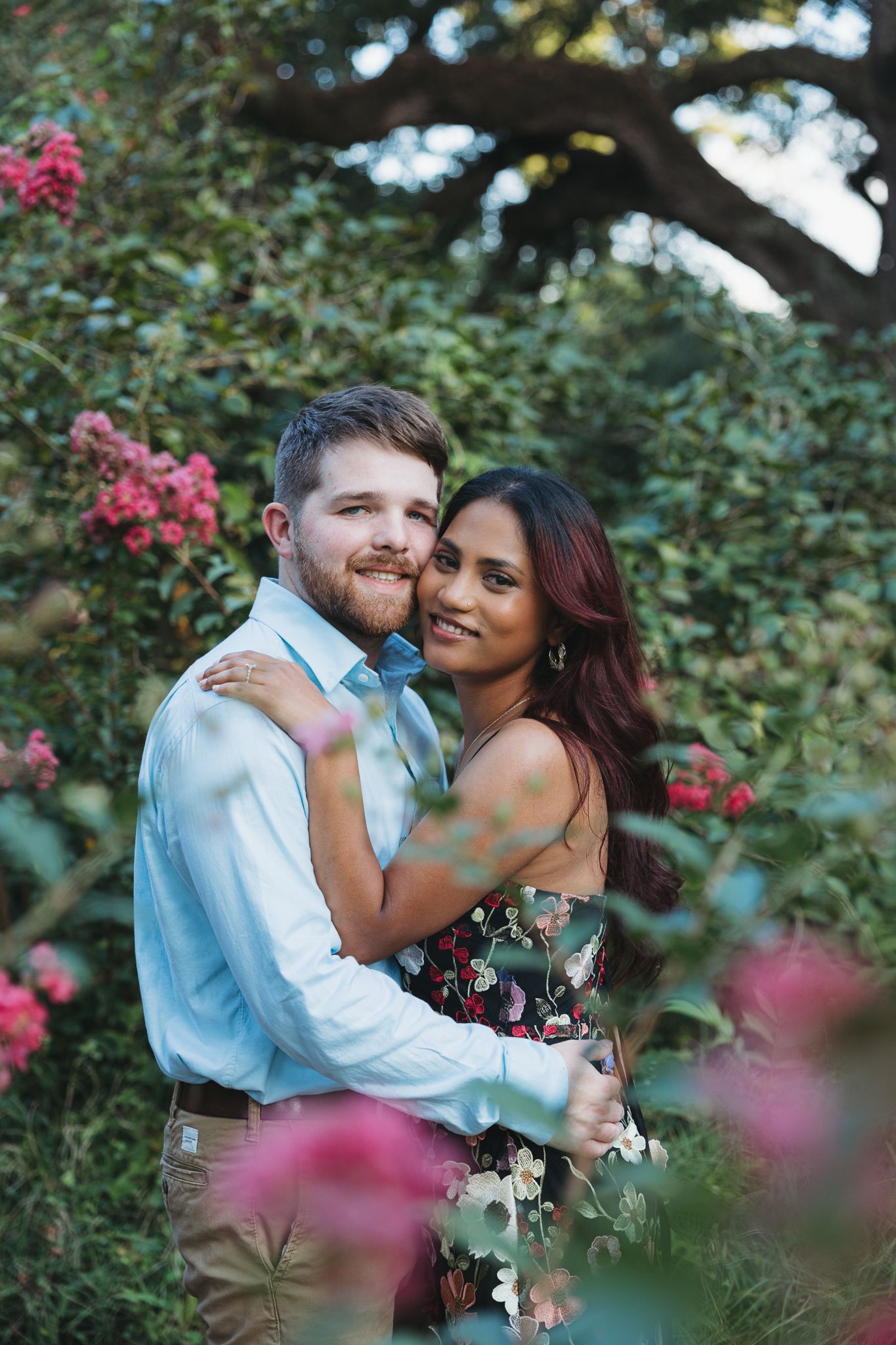 A happy couple embraces among lush green bushes and pink blooming flowers outdoors.
