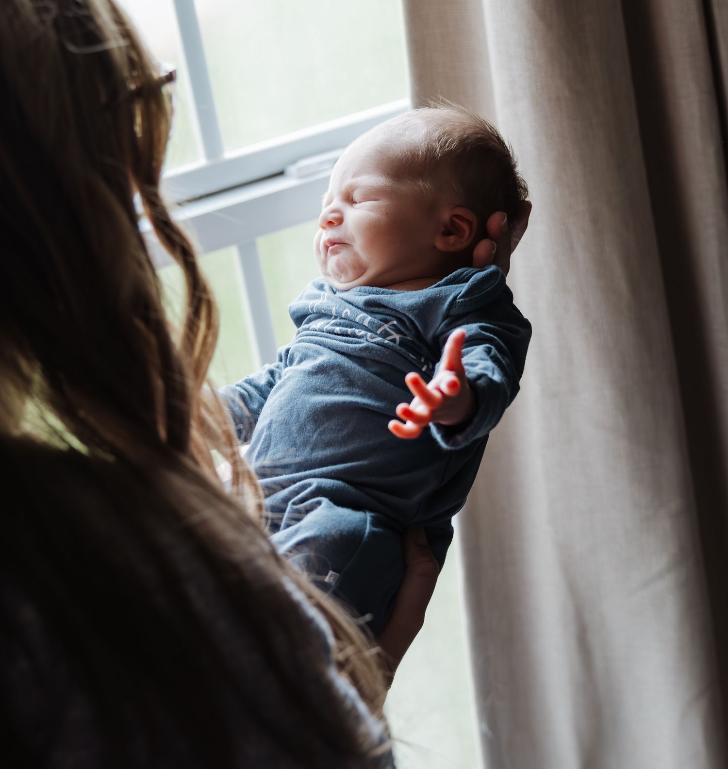 Close-up of sleeping newborn baby in blue outfit held near a sunlit window.