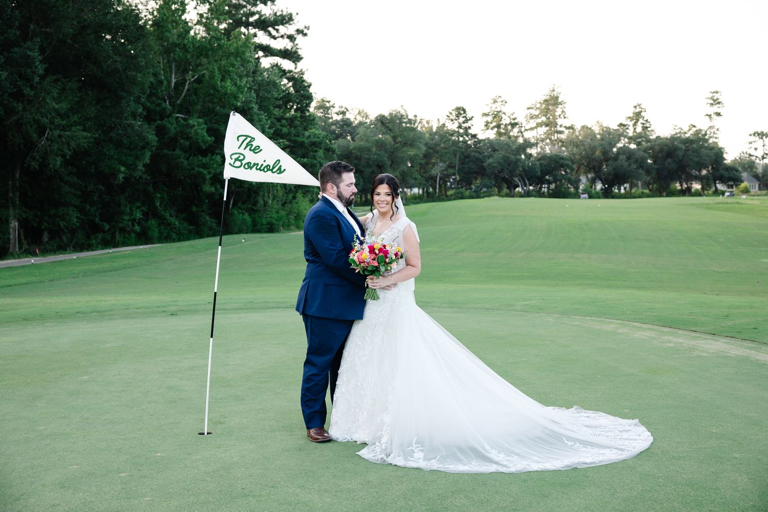 Bride and groom pose together on a lush green golf course beside a personalized flag on their wedding day.