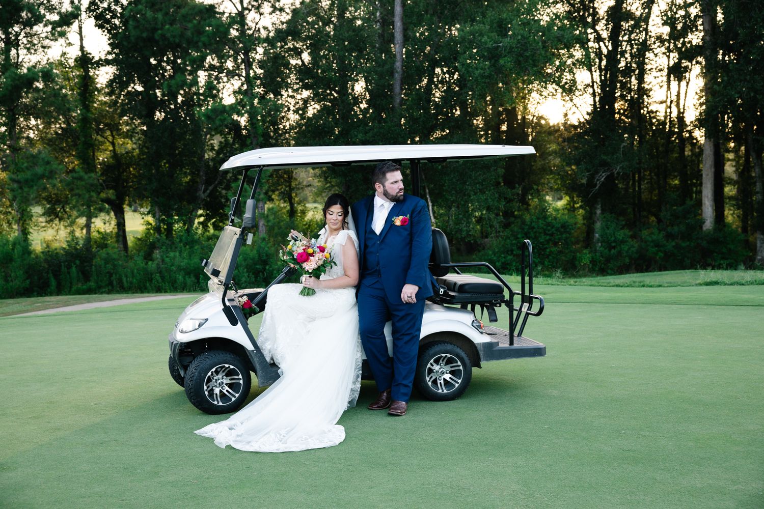 Bride and groom posing beside white golf cart on green golf course at sunset.
