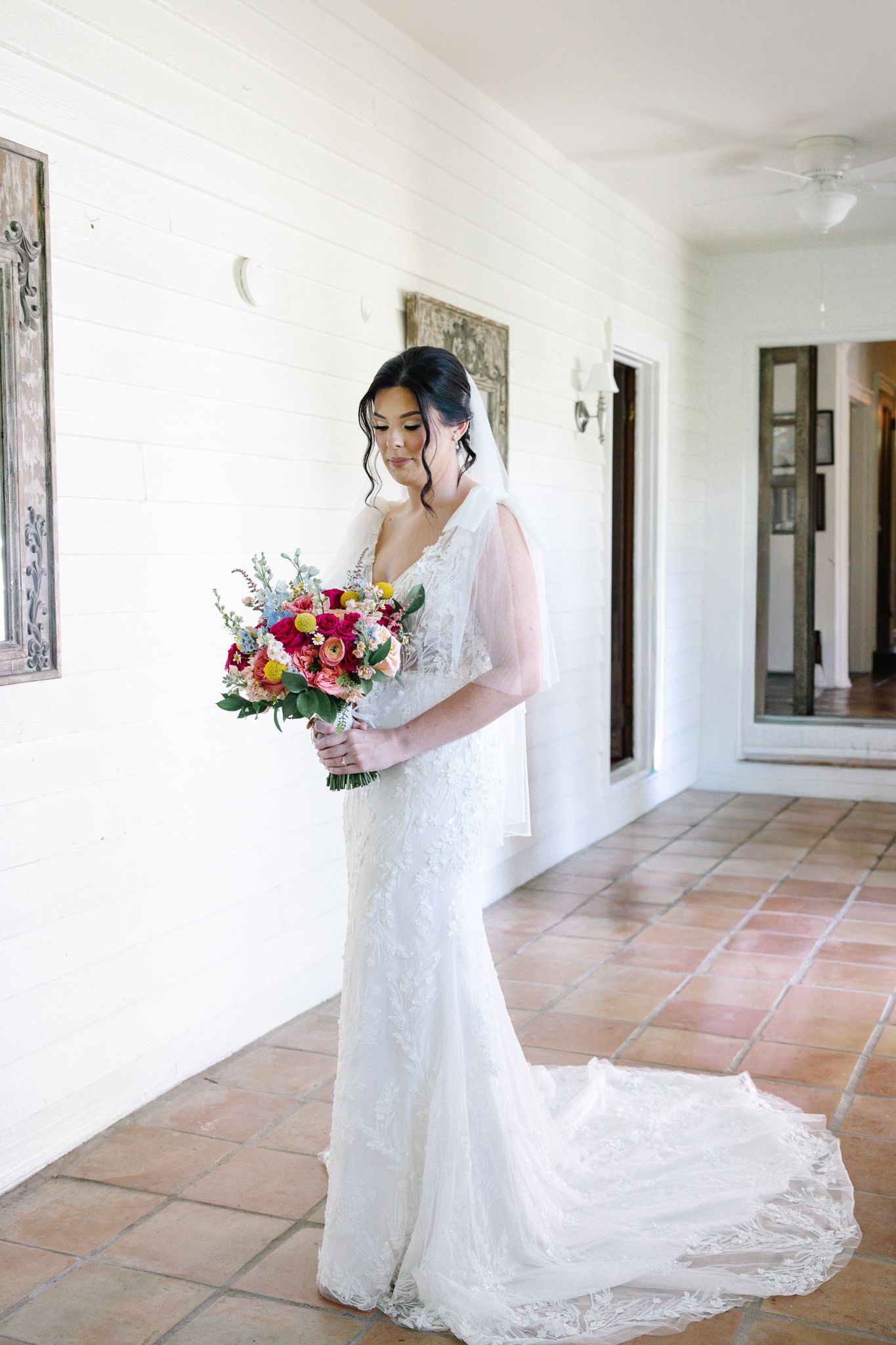 Elegant bride in fitted lace gown with long train holds vibrant bouquet while standing in a sunlit white corridor.