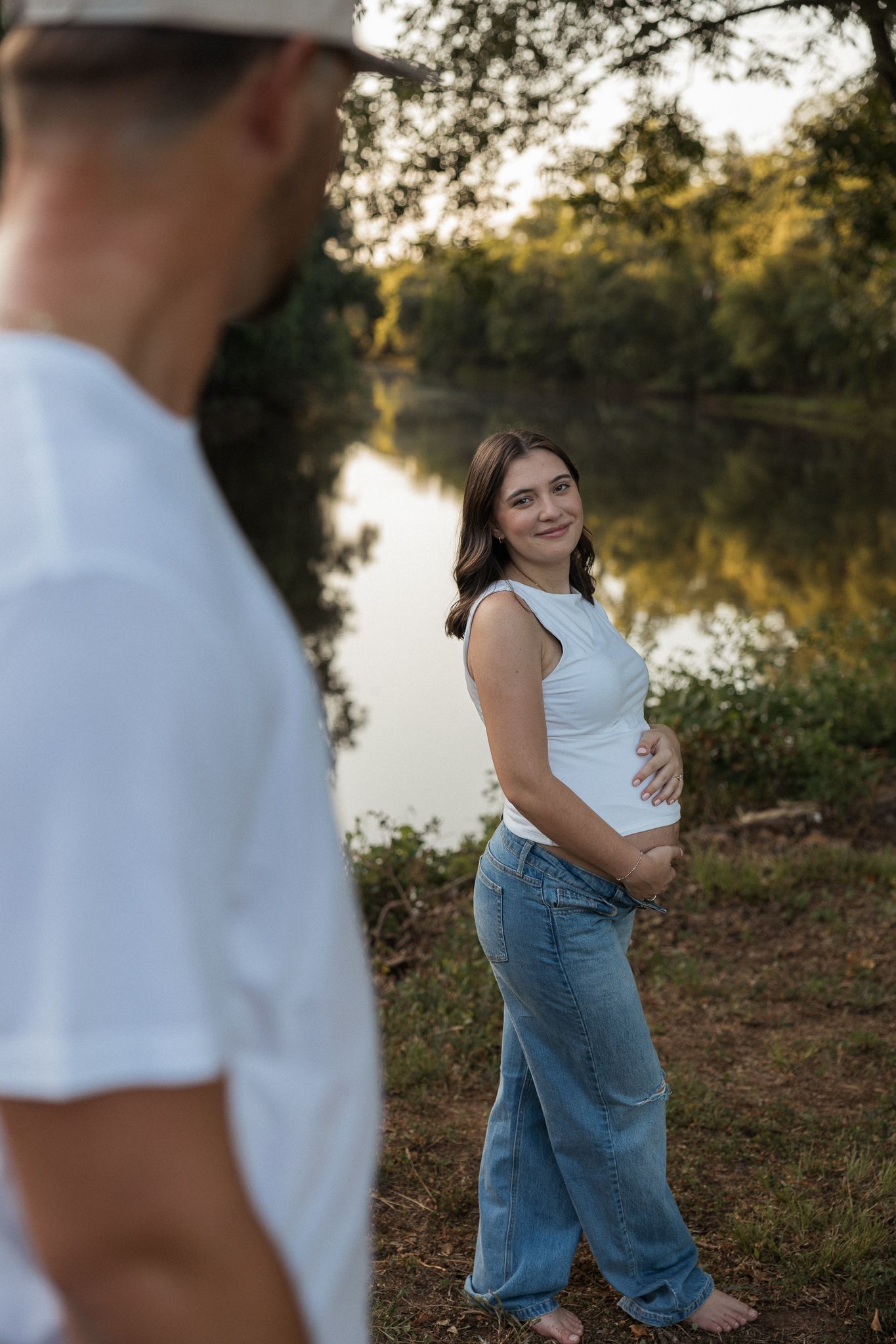 Smiling pregnant woman in white tank top and jeans holding belly near reflective lake at sunset.