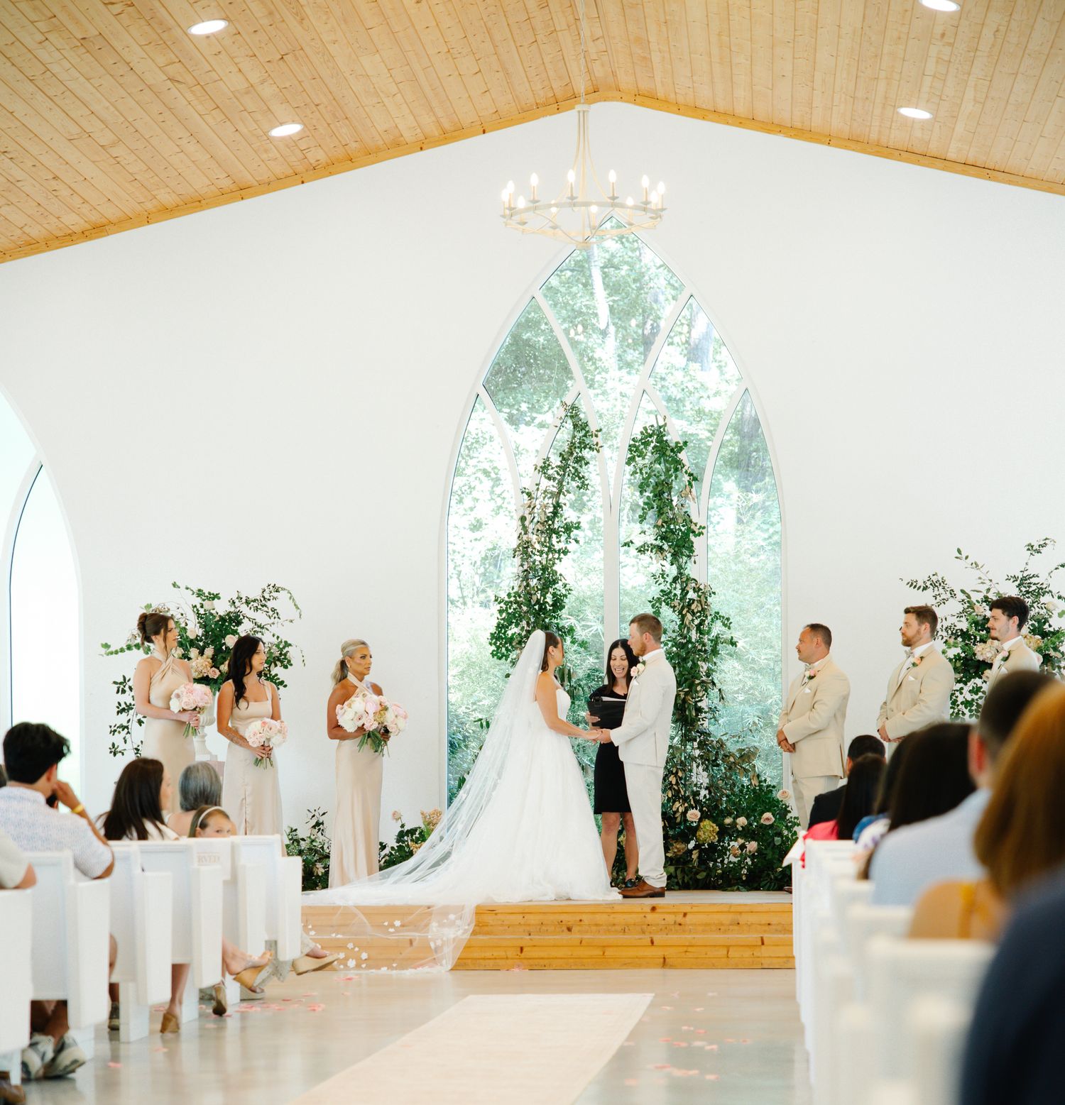 Elegant white chapel wedding ceremony with arched window, floral greenery arch, and guests seated in pews.