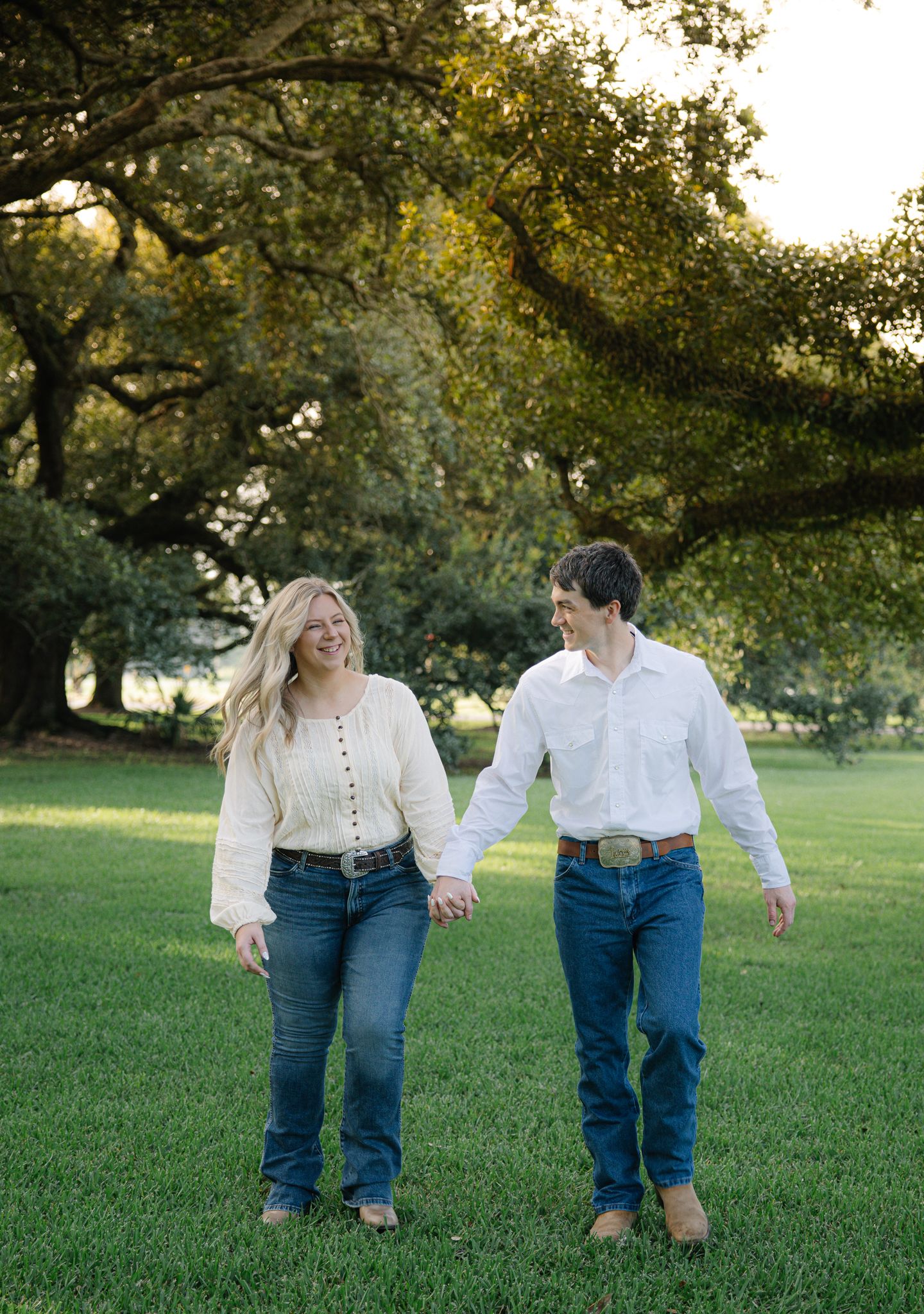 Happy engaged couple holding hands and walking through green park with large oak trees.